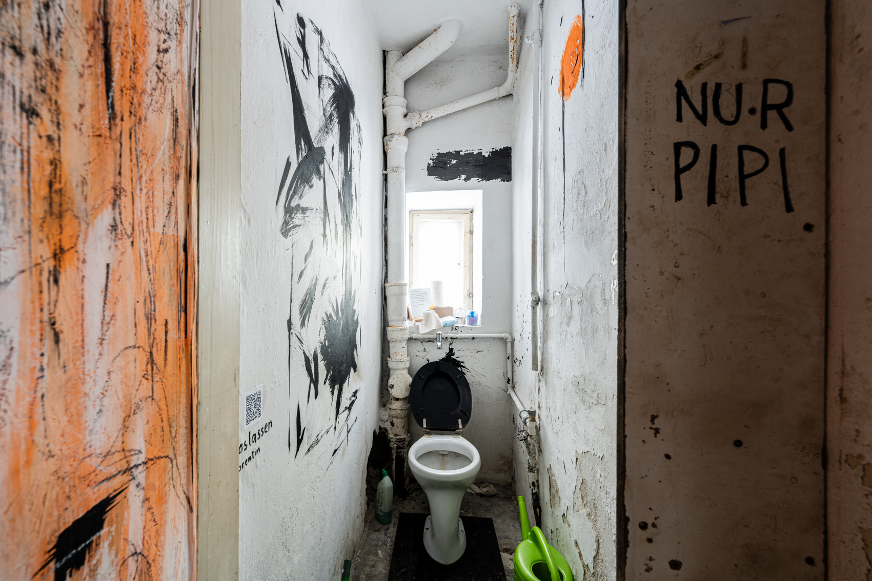 Bild 1 Loslassen: Ein schmaler Raum mit einem kleinen Fenster und einer Toilette darin. Der Klodeckel ist schwarz. An die linke Wand sind mit schwarzer Farbe kr&auml;ftige Pinselstriche gemalt. An der rechten Wand ist ein orangener fu&szlig;ballgro&szlig;er Kreis. Das Innere der T&uuml;r ist ebenfalls orange mit schwarzen schnellen Strichen bemalt.