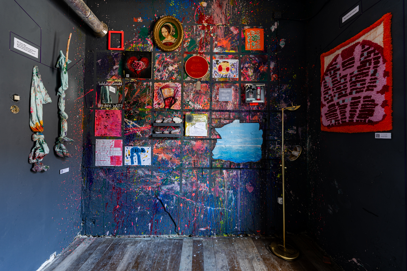 Bild 2 zu Mehr Raum f&uuml;r Wut:  Eine graue Wand des Raumes ist voller bunter Farbspritzer. Einzelne Gem&auml;lde und Gegenst&auml;nde zum Thema Wut h&auml;ngen ebenfalls an der Wand.