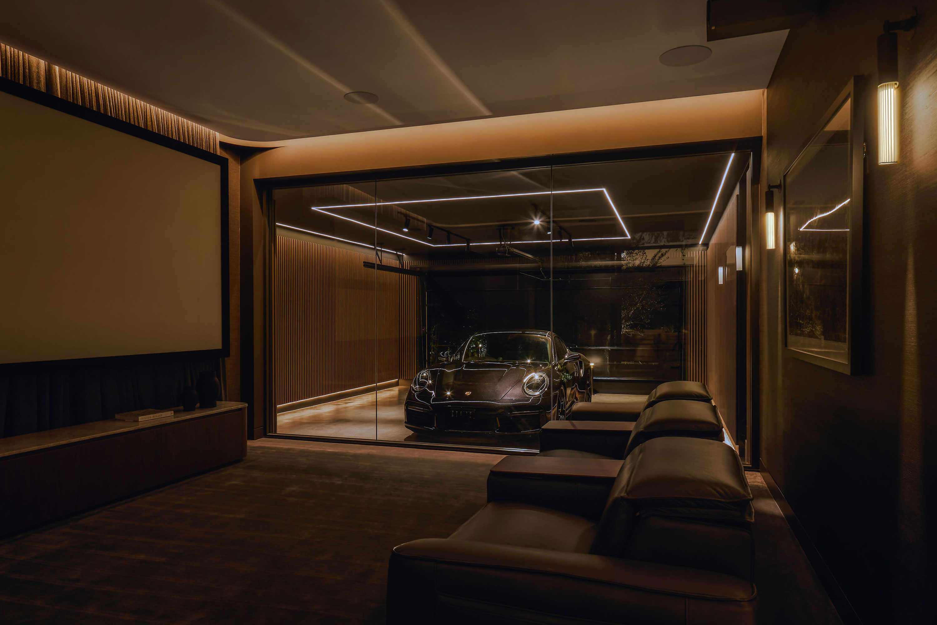 Luxury home theater room with leather seating and large screen, featuring a modern glass wall overlooking a high-end sports car in a sleek, well-lit garage with contemporary wood paneling and ambient LED lighting.
