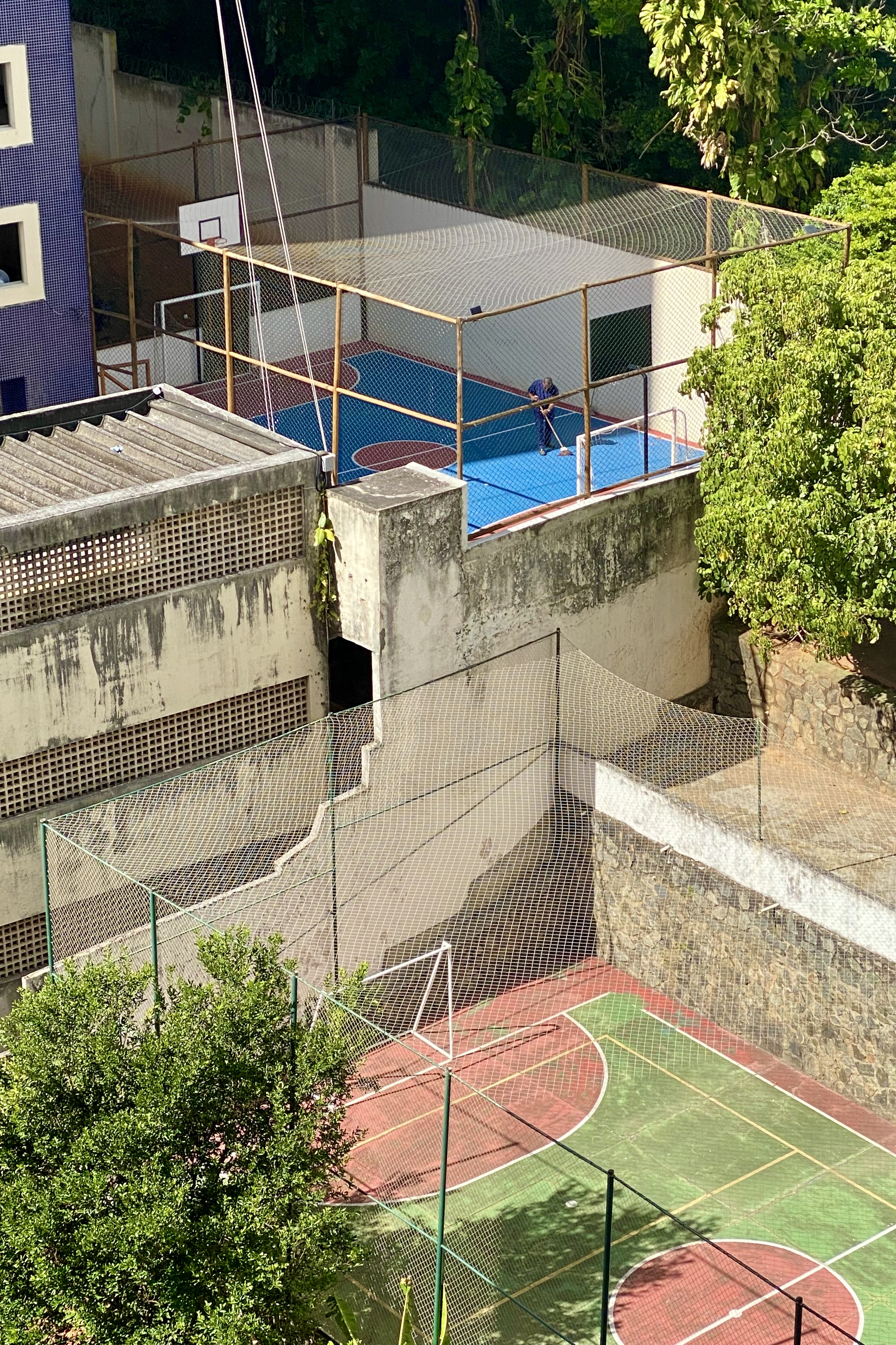 Rooftop sports area with blue basketball court and soccer field surrounded by nets, lush green trees in the background, and a sunny atmosphere in Salvador, Bahia, Brazil.