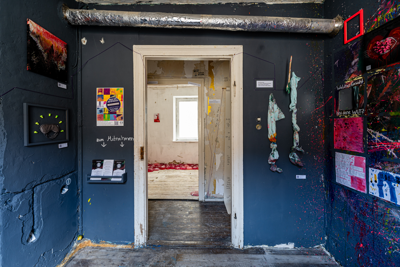 Bild 4 zu Mehr Raum f&uuml;r Wut: Aufnahme im Raum mit Blick auf den Ausgang und in den Gang. Gegen&uuml;ber ist ein Teil des Raumes vom Raum RED zu sehen. Links des T&uuml;rstocks befinden sich einige Informationsflyer zum Mehrfrau Kollektiv.