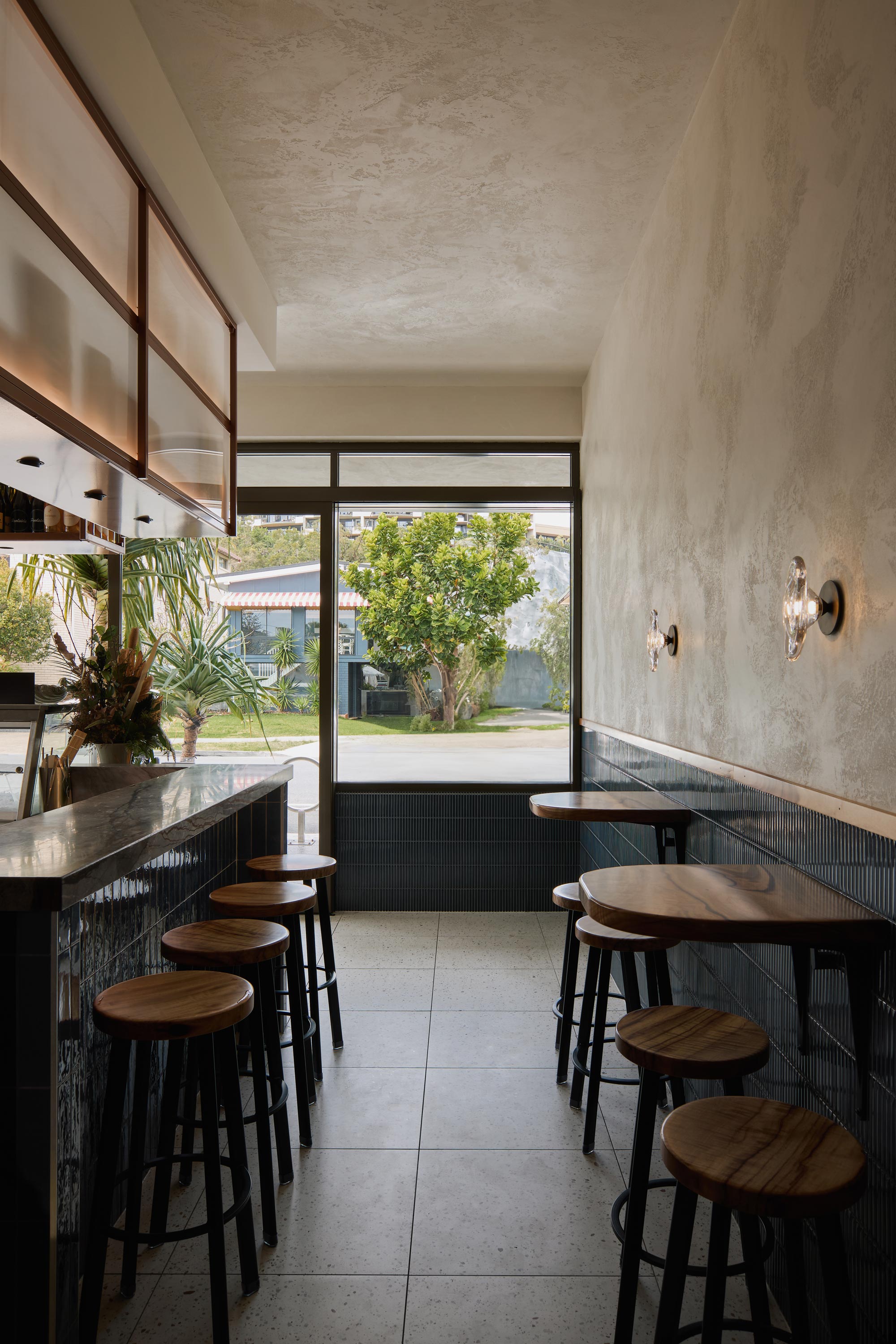 The material palette of a restaurant interior showcases richly textured stone surfaces, deep blue tiling and timber accents, creating an earthy elegance that resonates with Eska's oceanic theme. Large glass windows invite an abundance of natural light, connecting the interior space to the lush greenery outside.
