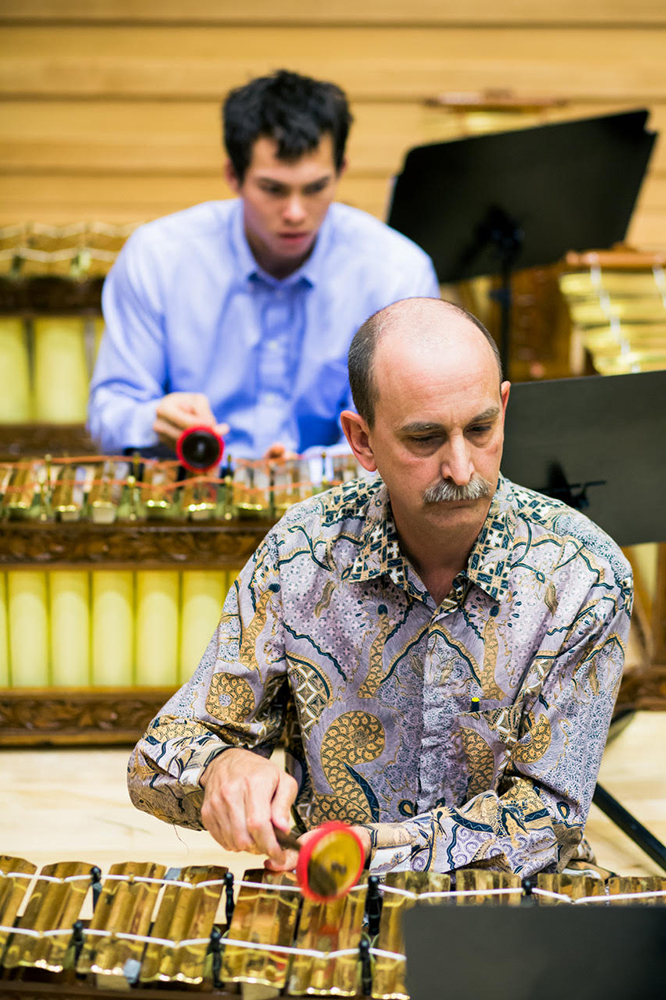 A cropped view of the musician on stage performing in the gamelan. One other musician, instruments, and music stands are also seen in the background.