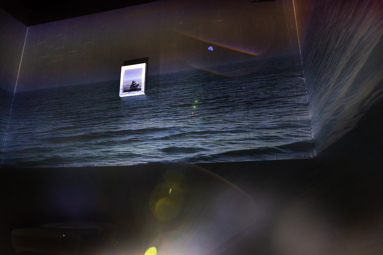 A dimly lit room-sized art installation where a projection of a seascape of dark blue water at night, is overhead the viewer and in the center of one wall is a small photograph with a white frame.