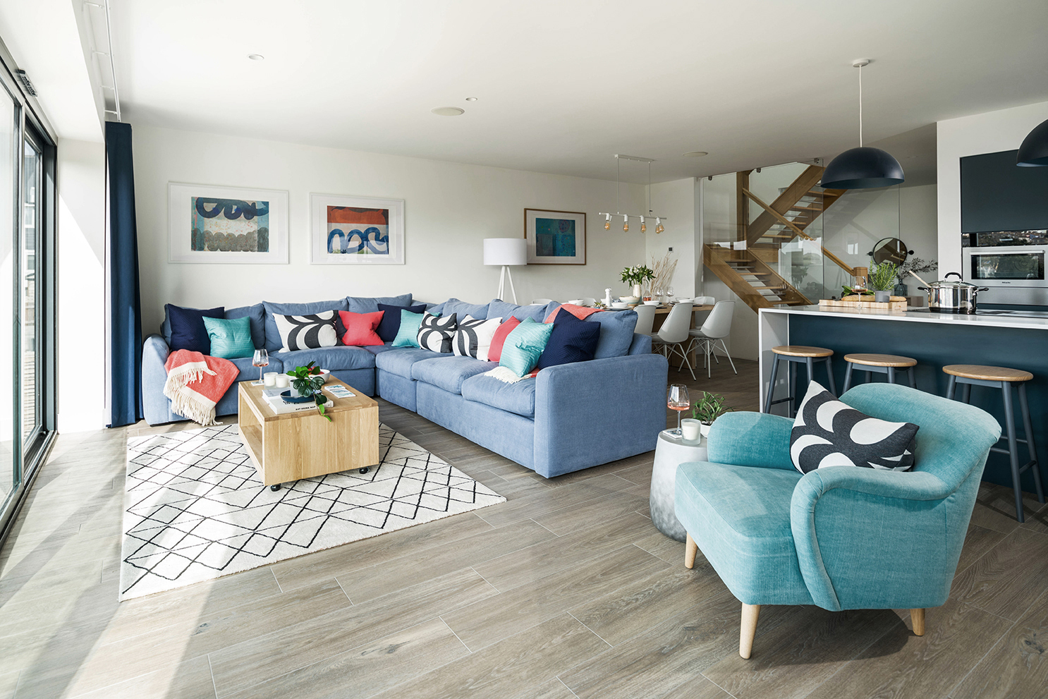 Modern, open plan lounge, dining room and kitchen. Blue and pink soft furnishings. Timber floor. Dark navy kitchen fixtures.