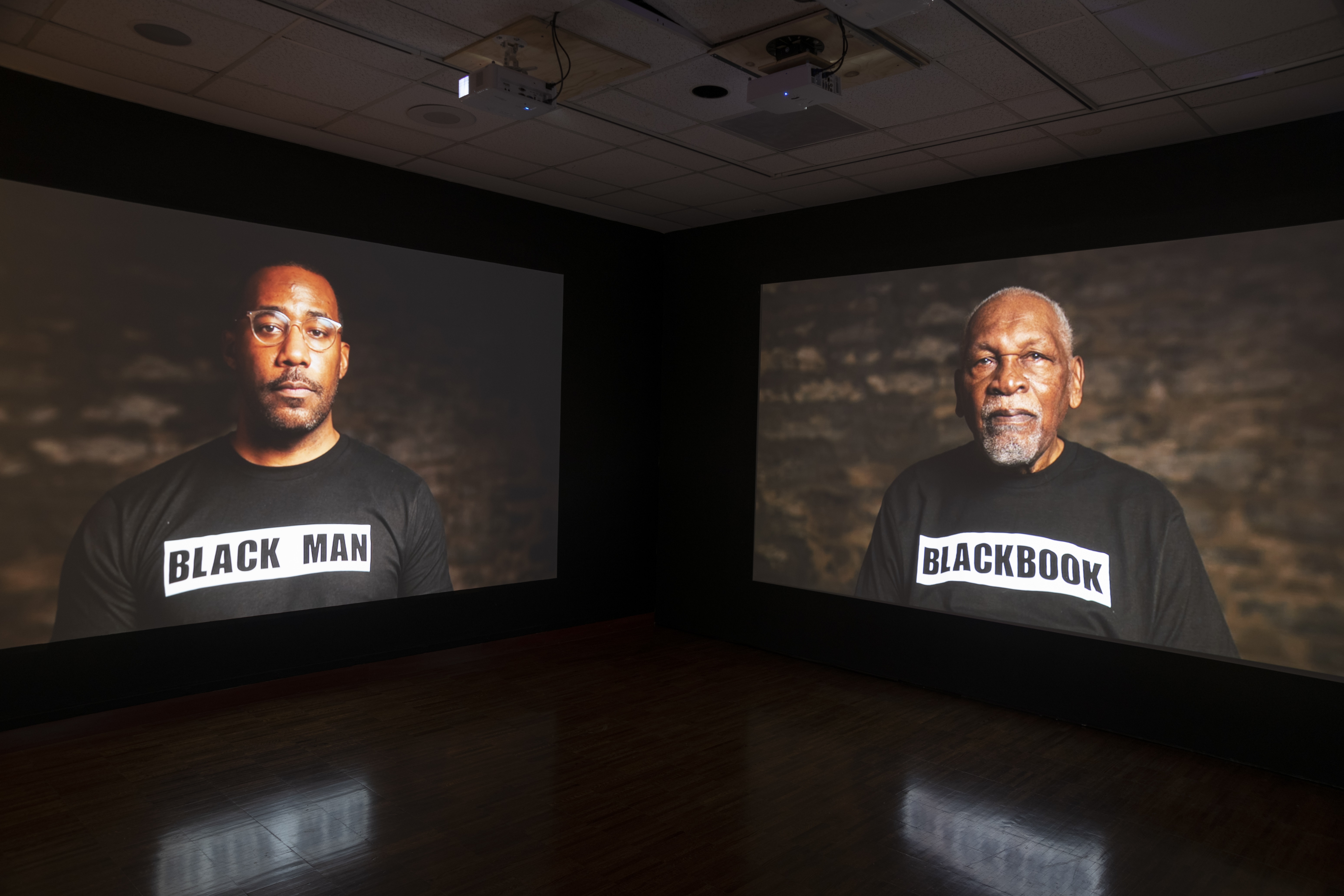 In a dark room, projectors display two different portraits on opposites walls of the same corner. The figure on the left is wearing a black t-shirt that reads “BLACK MAN” and the figure on the right is wearing the same, but reads “BLACKBOOK”