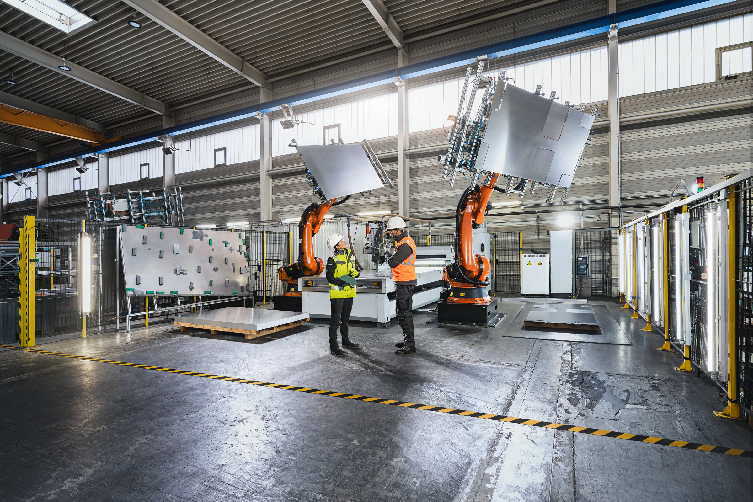 Ein Mann und eine Frau mit Sicherheitskleidung stehen in einer Fertigungshalle mit Industrierobotern. Kampagnenmotiv fotografiert von Peter Heck für die Firma DEKRA.