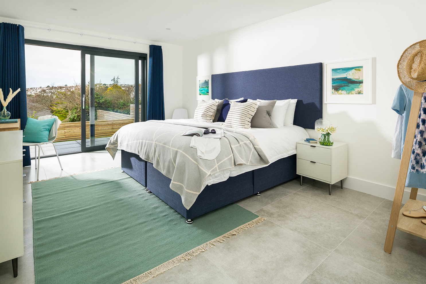 Bright, modern bedroom with double bed and clothing storage. Sea green rug on tiled floor. Door to balcony is open.