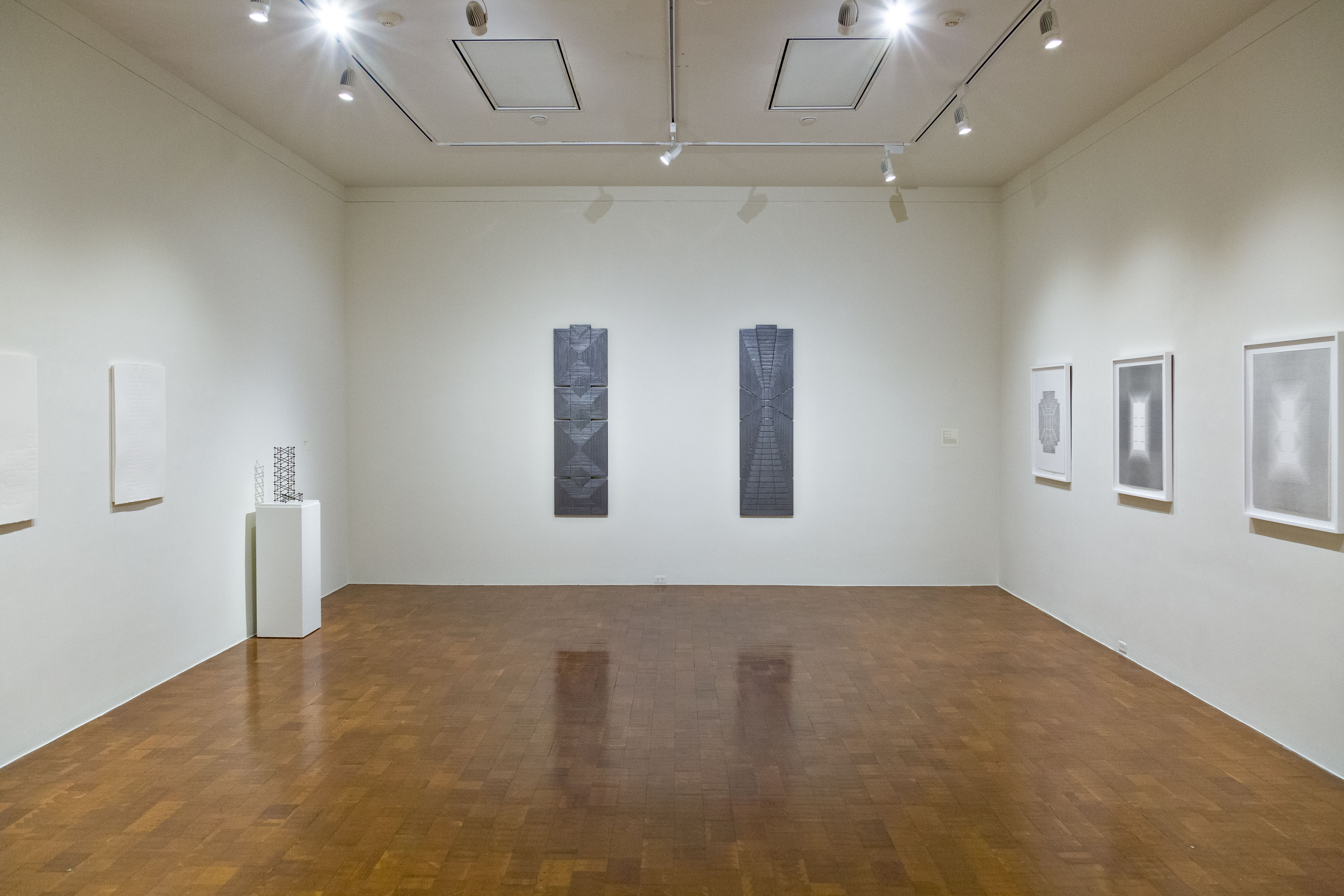 View of a sparse, white wall museum space with shiny wood floors and track lighting overhead. On the back wall, directly in front, hangs two long, rectangular dark grey and blue works. On the left and right walls to the side are more works, lighter in color, some framed, some unframed.