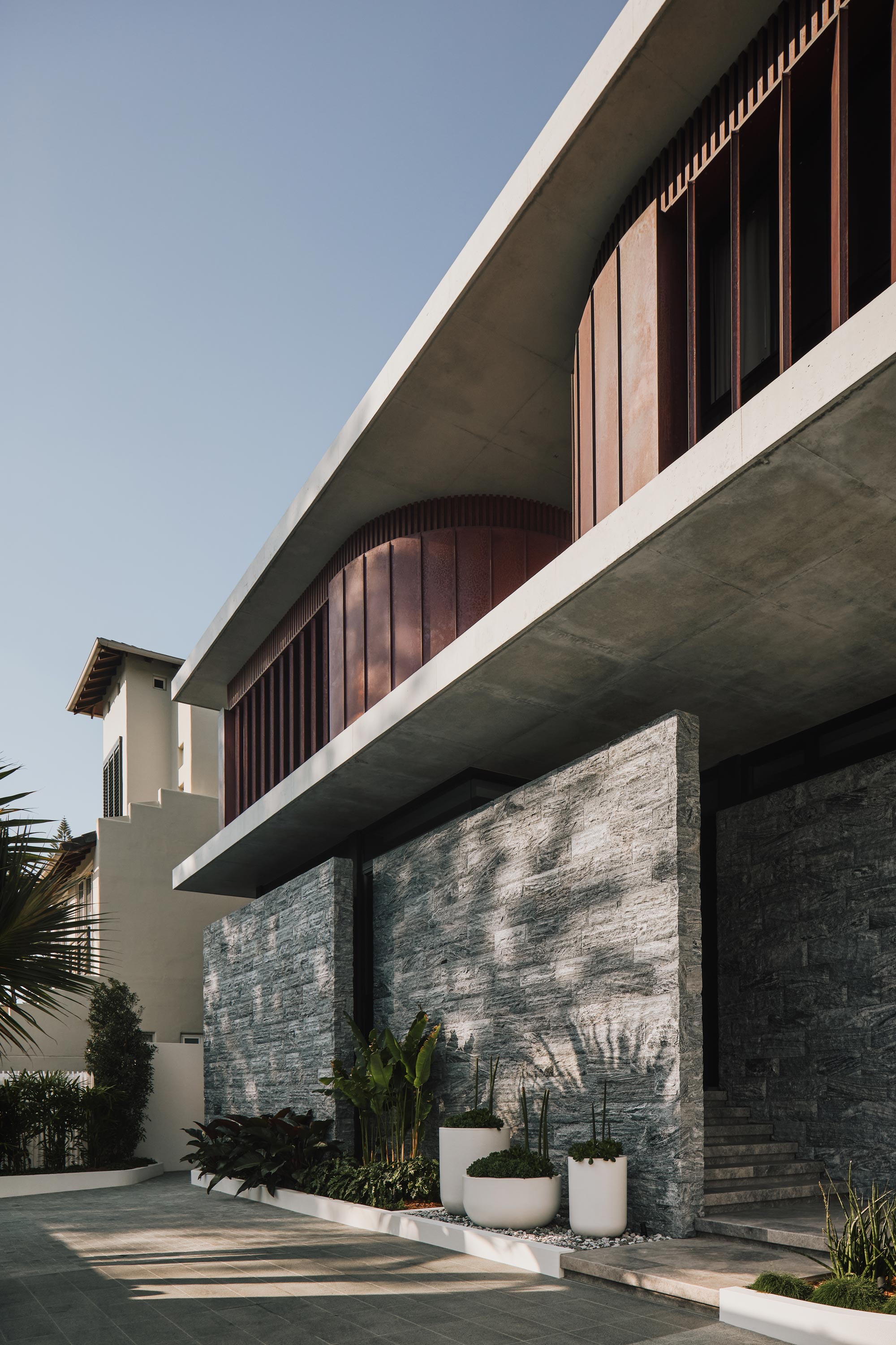 Close-up of the residence’s cantilevered concrete upper level with curved copper-clad facade, vertical timber battens, and textured stone-clad lower walls. Foreground features minimalist tropical planting in white pots.