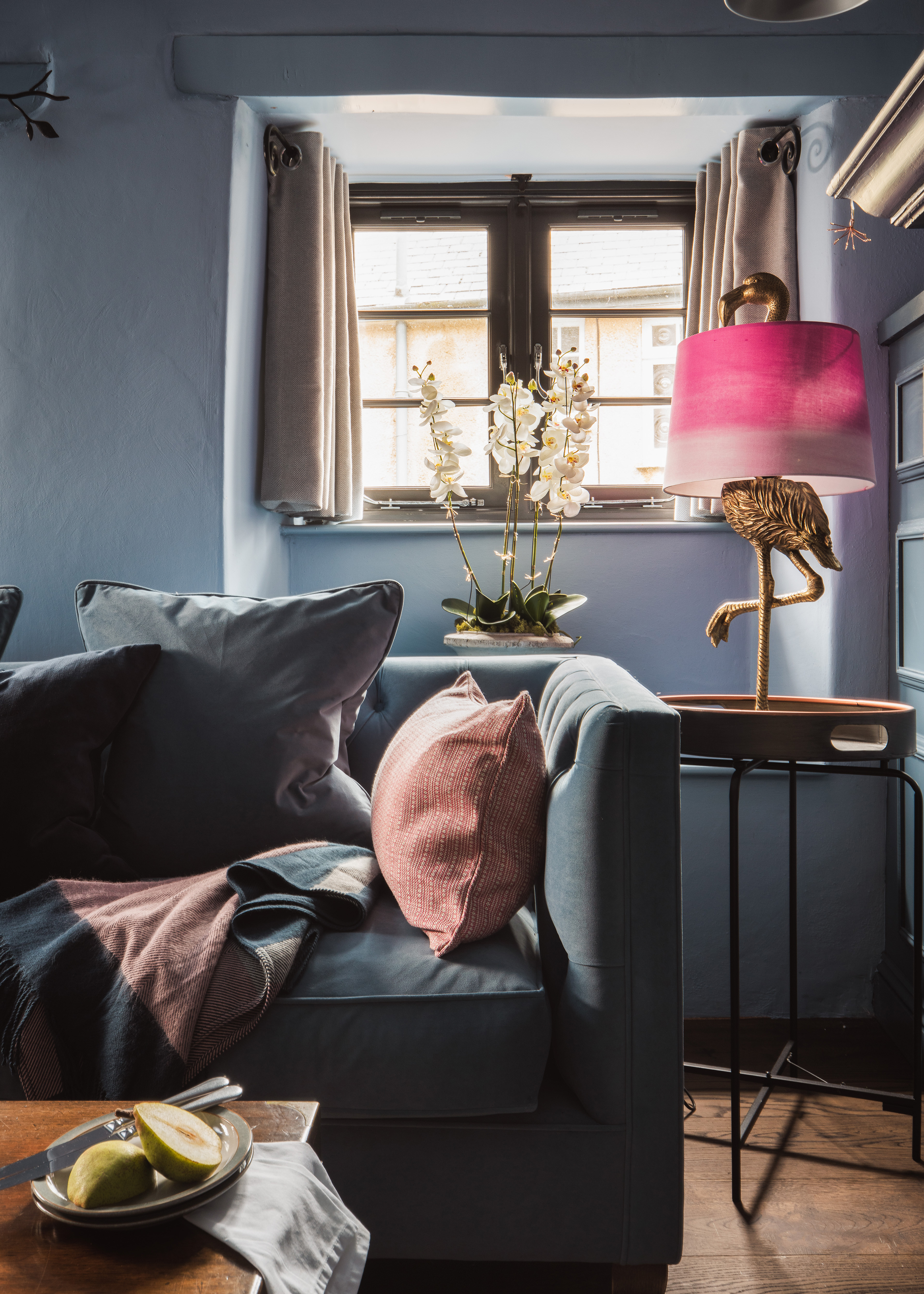 Close up of lounge with blue sofa, pink scatter cushion and ornate table lamp with flamingo stand and pink shade.