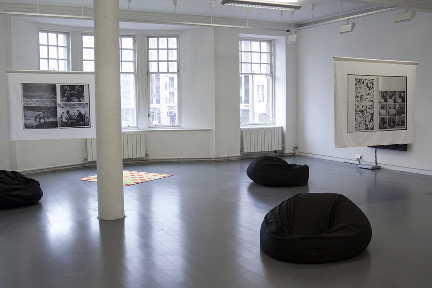 The image shows a mid-shot of the show After Hours, in a white gallery space with a grey floor and windows on the back wall. There are three black bean bags dotted around the floor, and towards the back there is a small patchwork quilt in high-visibility orange and yellow fabric. Two printed fabric banners hang from the ceiling, showing scanned double page spreads from the catalogue for the landmark 1970 exhibition 'Information' at MOMA, NY.