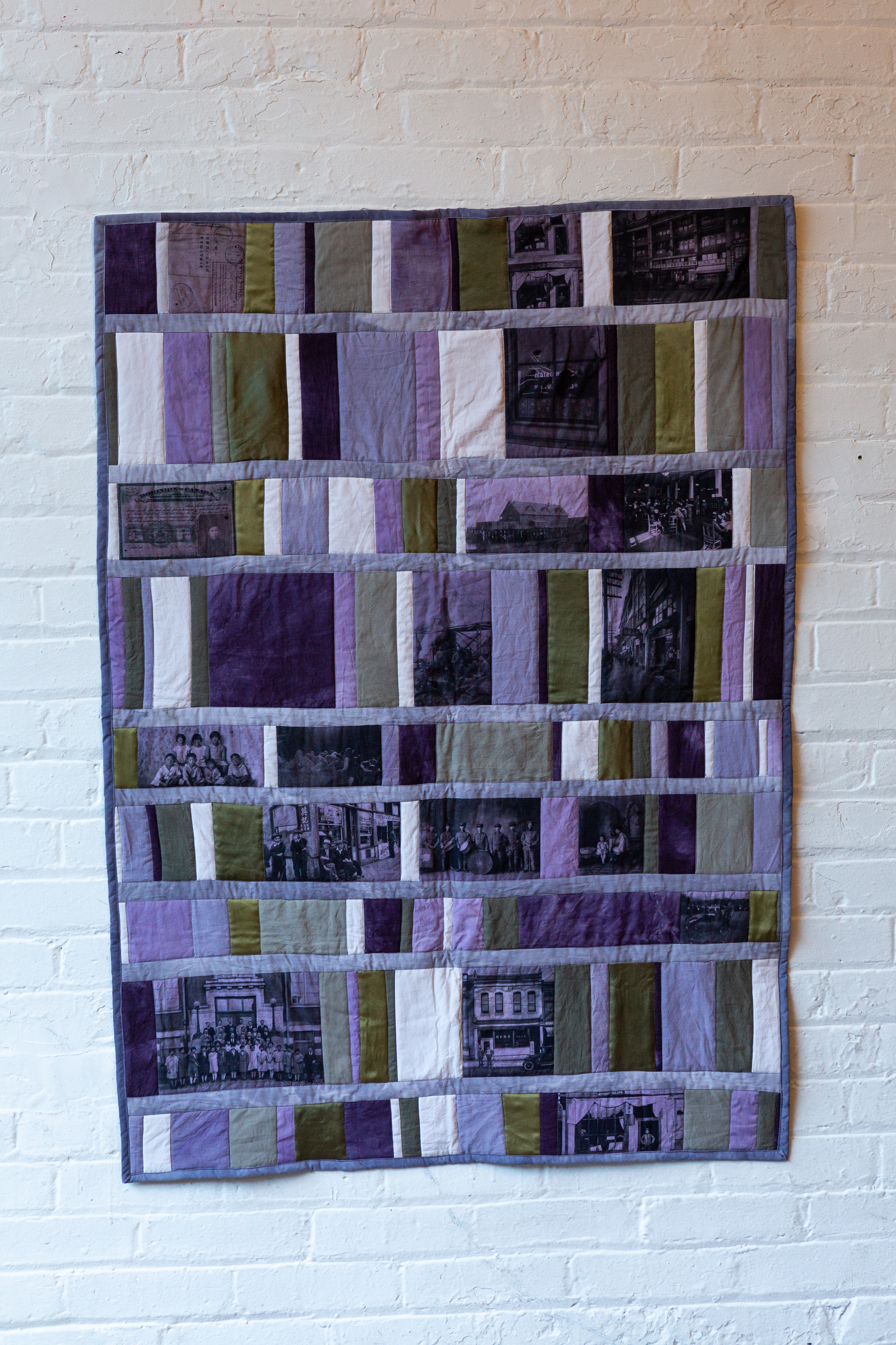A quilt of purple panels (light, medium, dark purple), white panels, and olive green panels. On several of the panels are black and white images of urban scenes and groups photos of Chinese Canadians, both adults and children rendered with a purple-y hue.