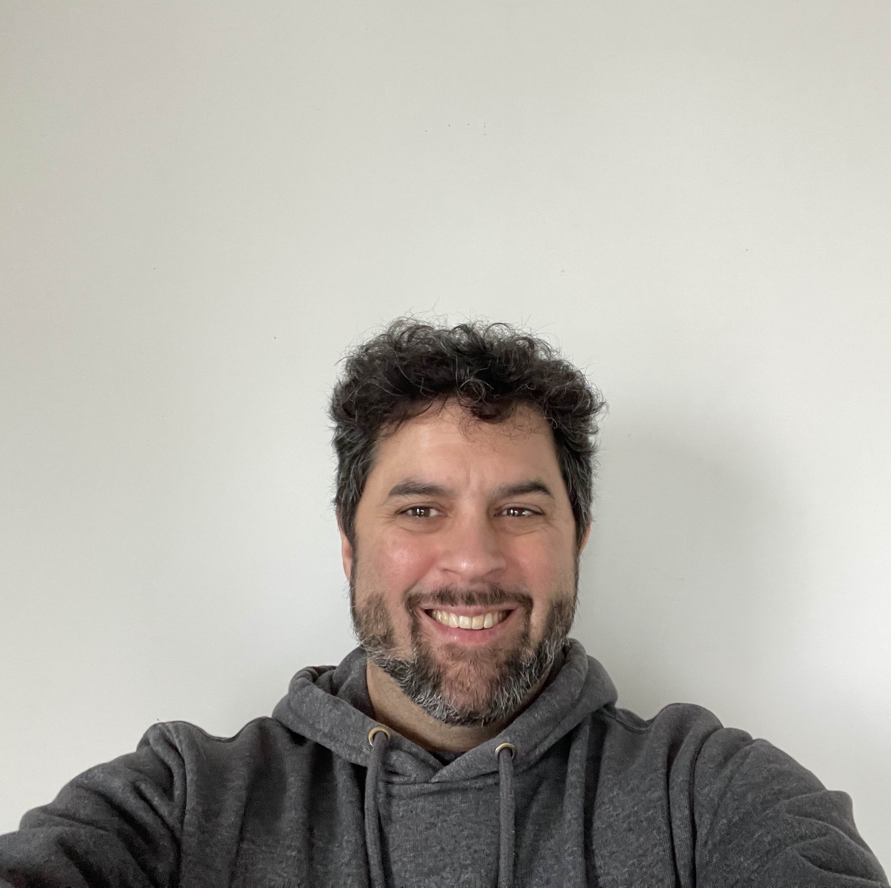 Person wearing a grey hoodie sweatshirt. They have a grey beard, mustache, and short curly hair. They are smiling and looking slightly to the right.