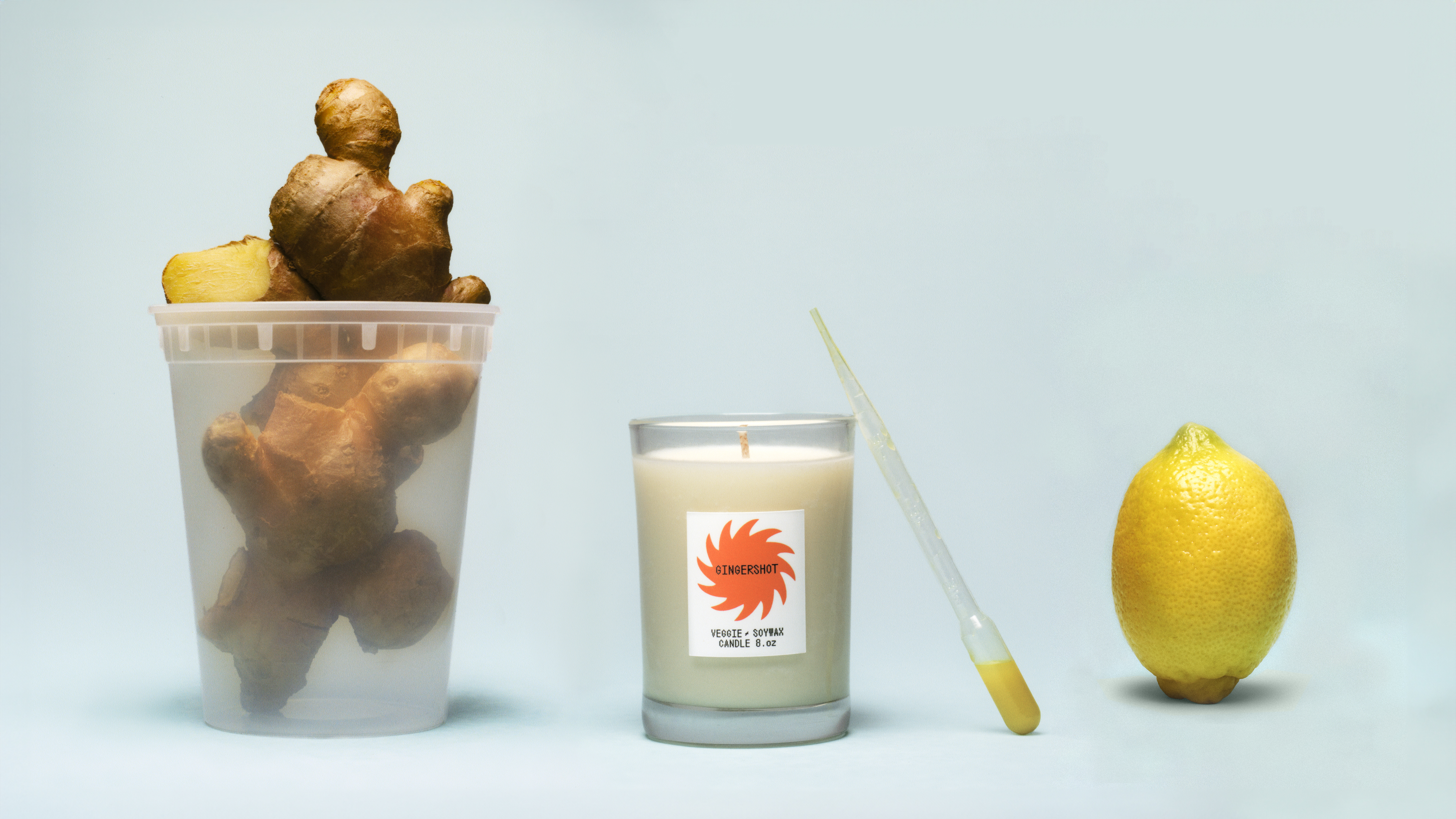 A cup of ginger root next to a candle with a pipet on it, next to a lemon