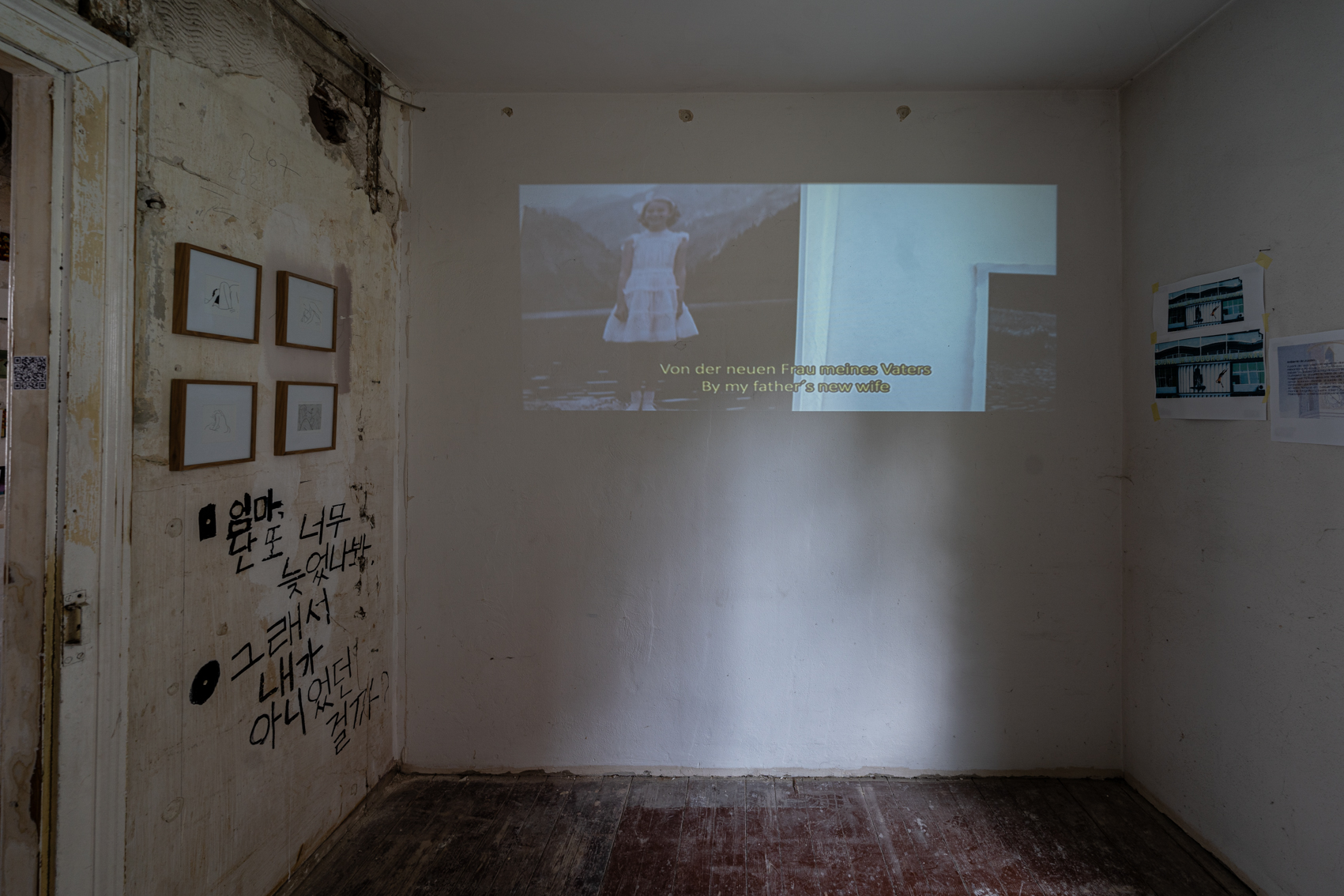 Bild 1 zu A la deriva: Ein Beamer projiziert an eine wei&szlig;e Wand einen Ausschnitt aus einem Film mit englischem und deutschem Untertitel. An der Wand links neben der T&uuml;r h&auml;ngen vier Zeichnungen in Bilderrahmen. Diese zeigen in vereinfachten Linien Intimit&auml;t zwischen zwei Personen.