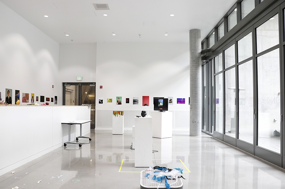 View of an exhibition in a sun-drenched gallery where printed photos hang on the walls and artworks of many mediums, including video and found objects, are installed throughout the space.