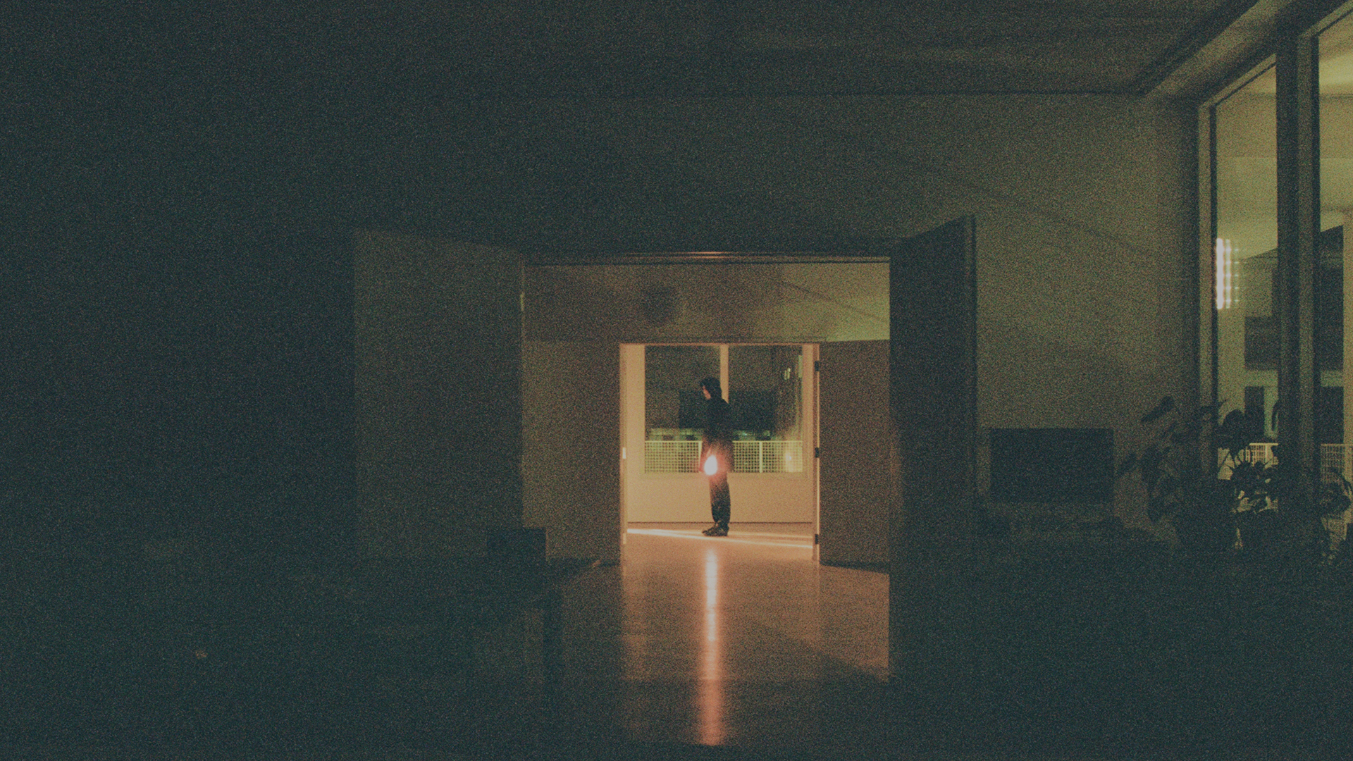 A grainy analogue photograph of a figure in the distance, through two open sets of doors in the centre, at the end of a series of empty rooms.