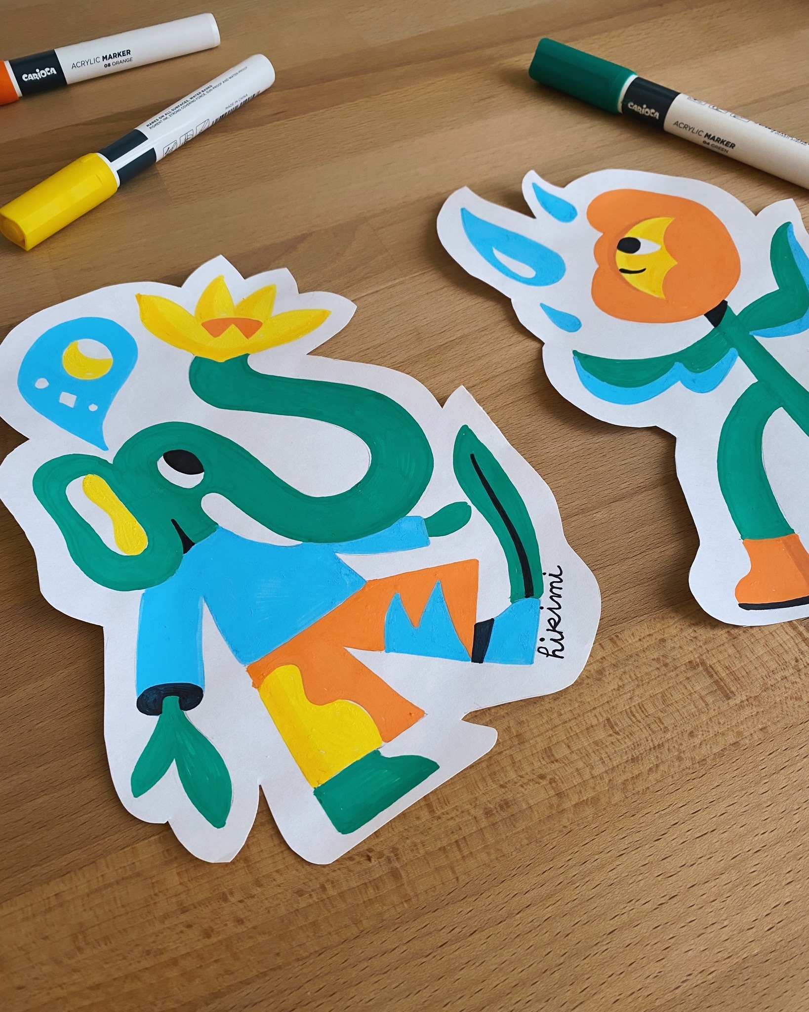 A close-up of two hand-painted cut-out illustrations on a wooden desk. One features a whimsical green elephant character with a yellow flower on its head, wearing colorful clothes. The other is a playful anthropomorphic flower with an orange face and blue water-like elements around it. Acrylic markers in yellow, white, and green are scattered nearby. The artist's signature, 'hikimi,' is visible on the elephant illustration.