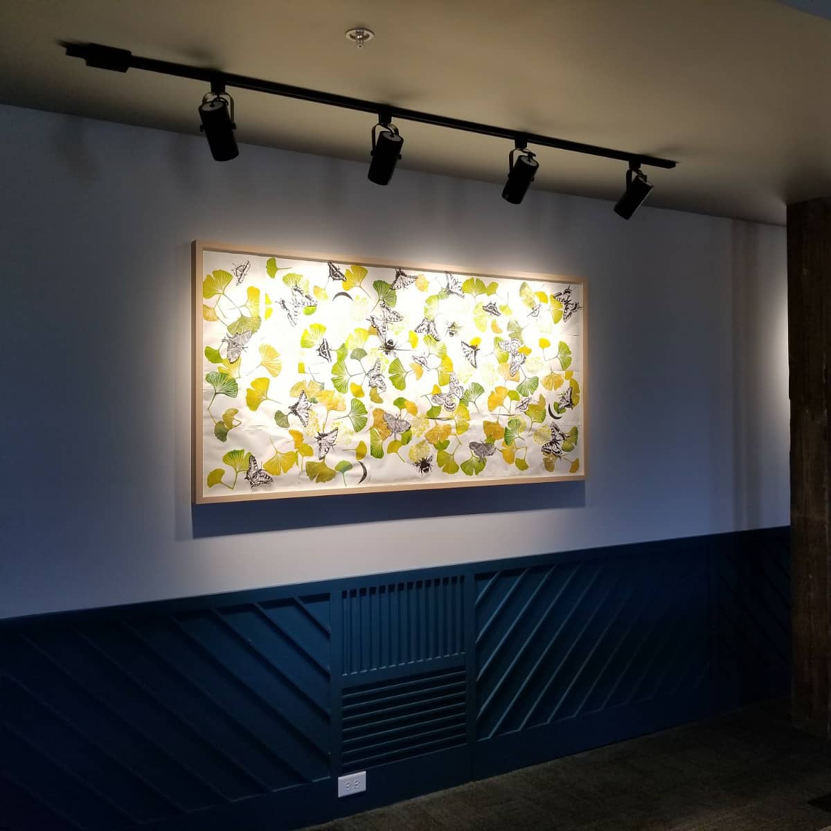 A dim gallery wall with a single, large print artwork showing an evenly spread array of ginkgo leaves in yellows greens, and browns on a white background.