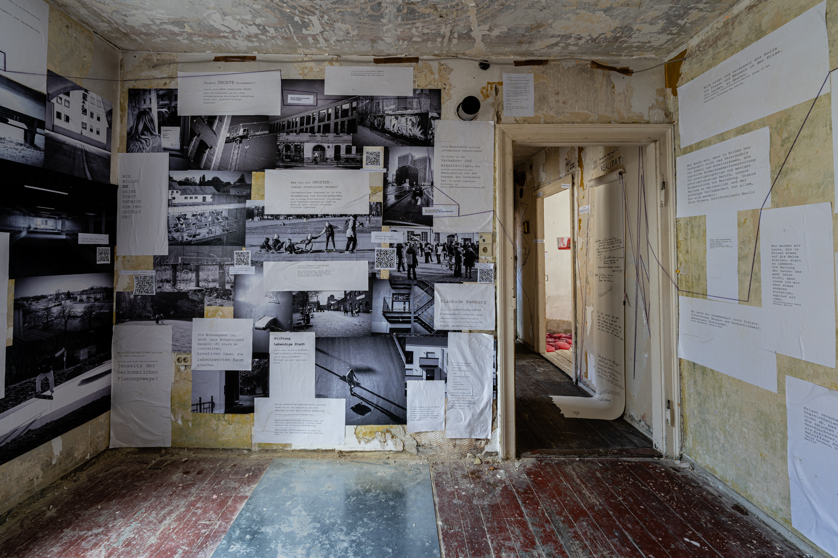 Bild 1 zu Die Bilder der Anderen: Blick im Raum stehend auf die Wand links der Eingangst&uuml;r. An der Wand eine Fotocollage aus vielen Schwarz-Wei&szlig;-Fotografien von St&auml;dten. Au&szlig;erdem Fragen und Antworten von Interviewten zum Thema Stadt auf wei&szlig;em Papier mit Computerschrift. 