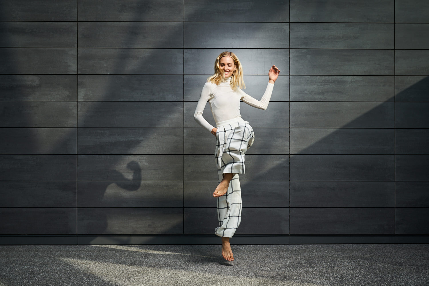 Eine hell gekleidete junge Frau hüpft vor einem grau gekachelten Hintergrund. Die Szene ist schoen, mit Lichtreflexen am Hintergrund, ausgeleuchtet.
