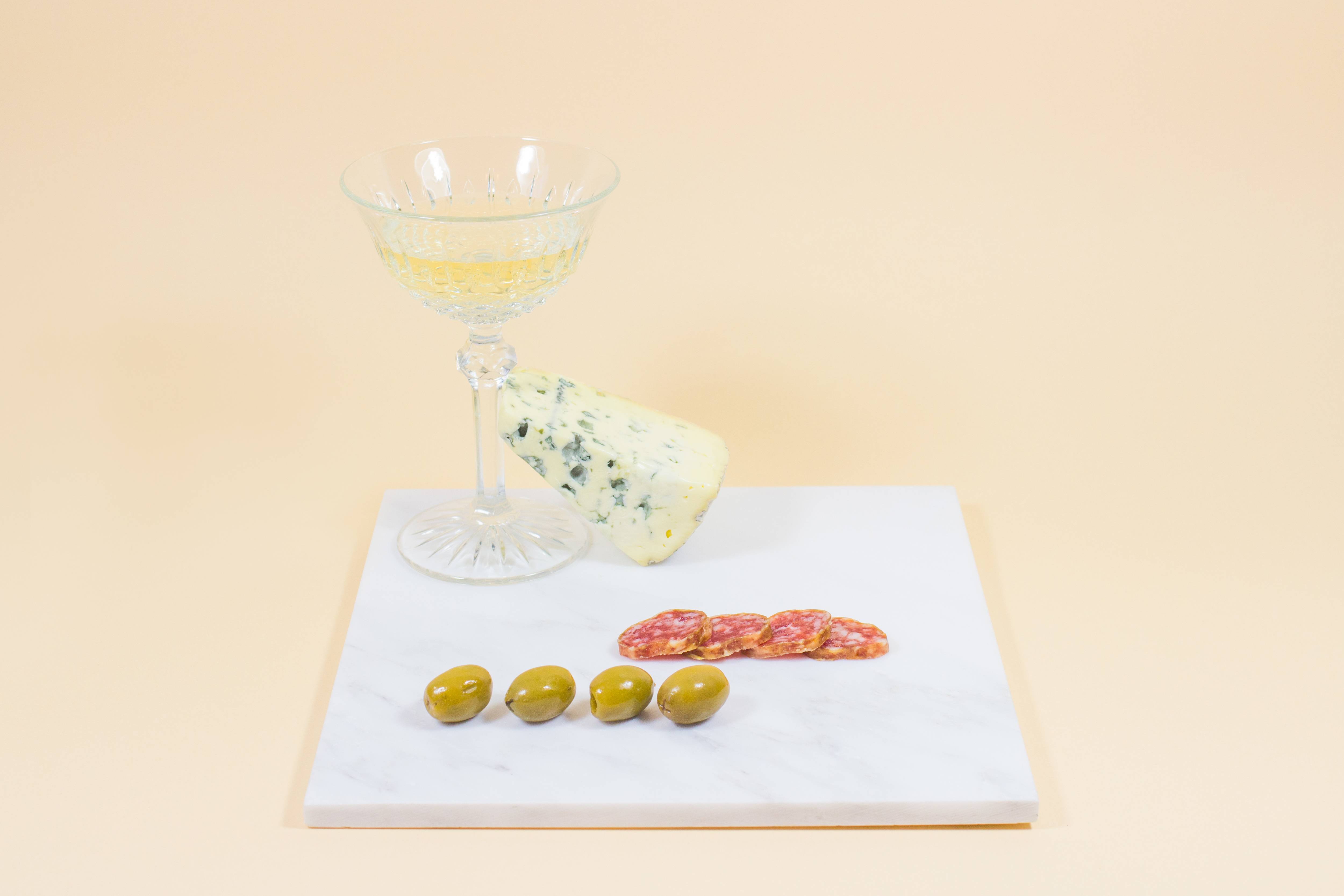A minimalist representation of an "Aperitivo" using olives, blue cheese, cured meat and white wine, in a beige set.