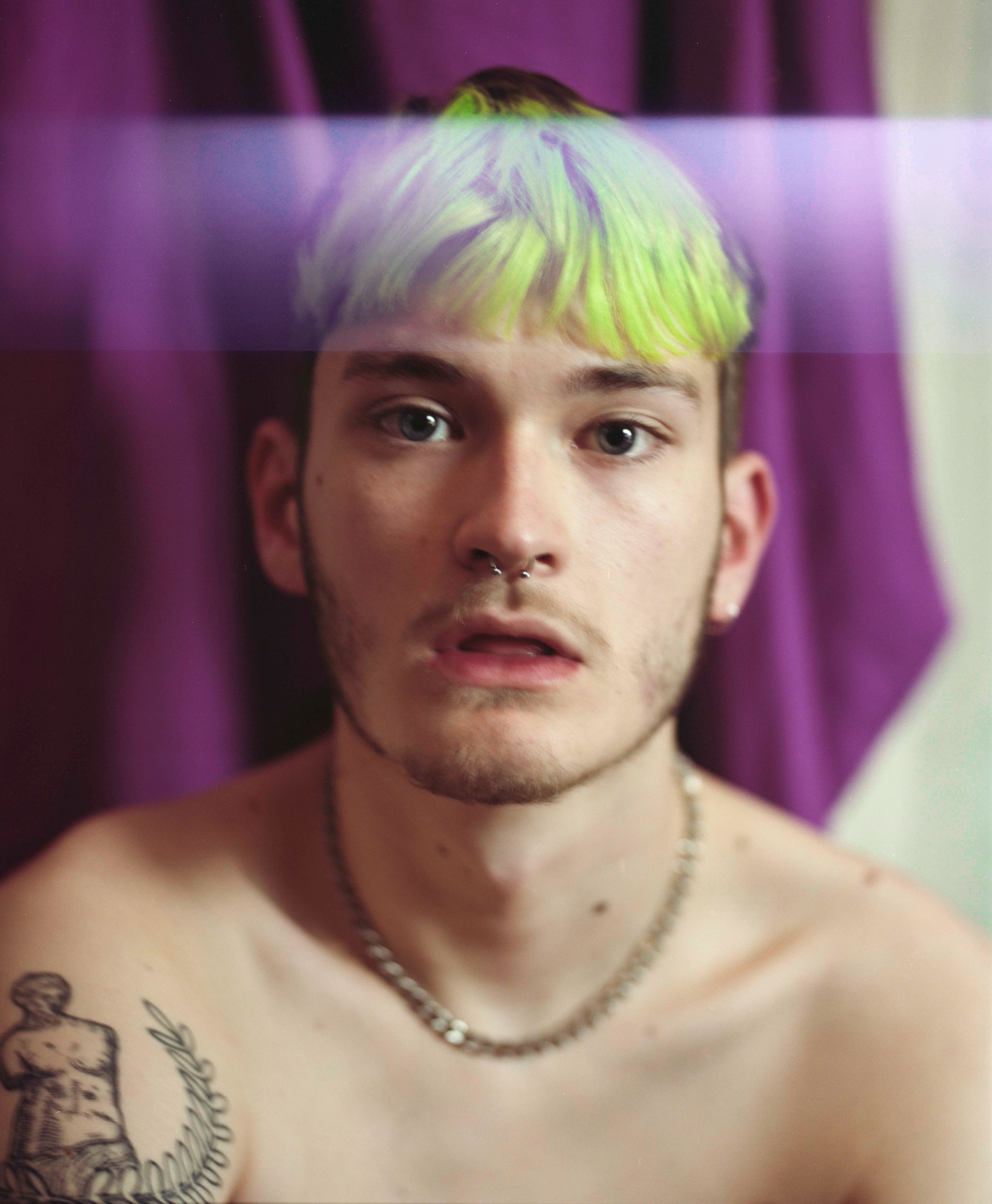 Portrait of boy with green hair. He has a shoulder tattoo and wears a silver chain