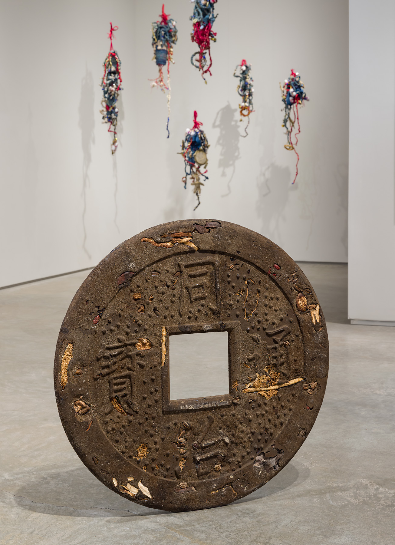 Front: Kenneth Tam, Why do you abuse me, 2022. Epoxy resin, dirt, sand, dried mushrooms, dried seaweed, dried bamboo shoots, dried jujube, dried goji berries, preserved apricots, sunﬂower seeds, dried roots, dried sweet potato, steel, 30 x 30 x 1 1/2 inches. Courtesy of the artist and Commonwealth and Council. Back: Elvira Clayton, Indigo Bess and her Children, 436 project, 2024. Six sculptures: indigo-dyed osnaburg, cotton, wire, jute, sequin, mini glass bottles, cotton bolls, shells, twigs, screen-printed archival text and photographs, embroidery hoops, acrylic, handwoven, and crochet. Bess: 37 x 12 x 7 inches; Sam: 46 x 12 x 5 inches; Elizabeth: 37 x 7 x 5 inches; Sena: 29 x 10 x 7 inches; Affa: 31 x 7 x 4 inches; Hannah: 37 x 10 x 6 inches. Courtesy of the artist. Photo by Etienne Frossard.