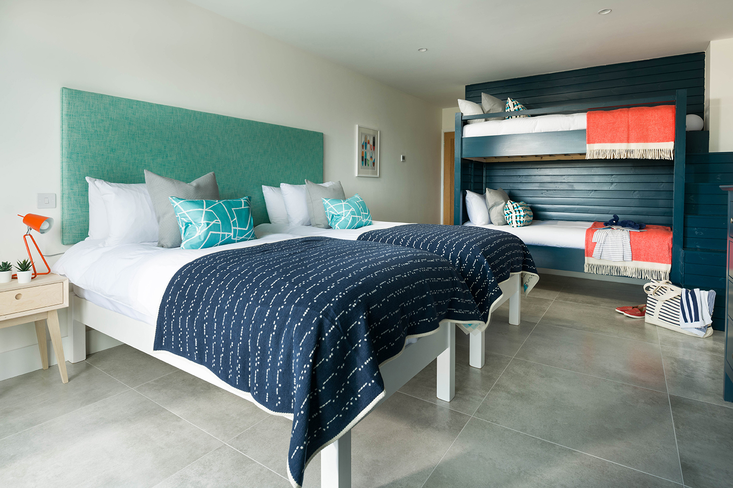 Modern bedroom with double bed and bunkbeds, and blue and red textiles. Tiled floor. Dark timber cladding and paint on walls.