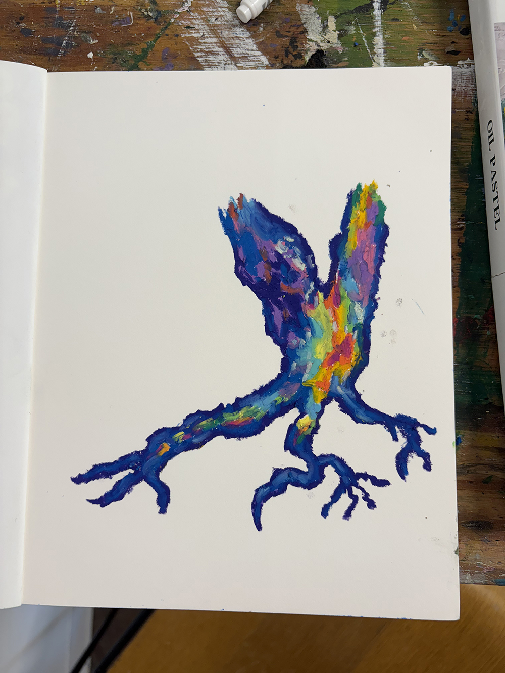 A drawing of what resembles tree trunks and roots in an open note or stretch book that is placed on a table. Partial view of an item, also on the table, shows a label that says "oil pastel".
