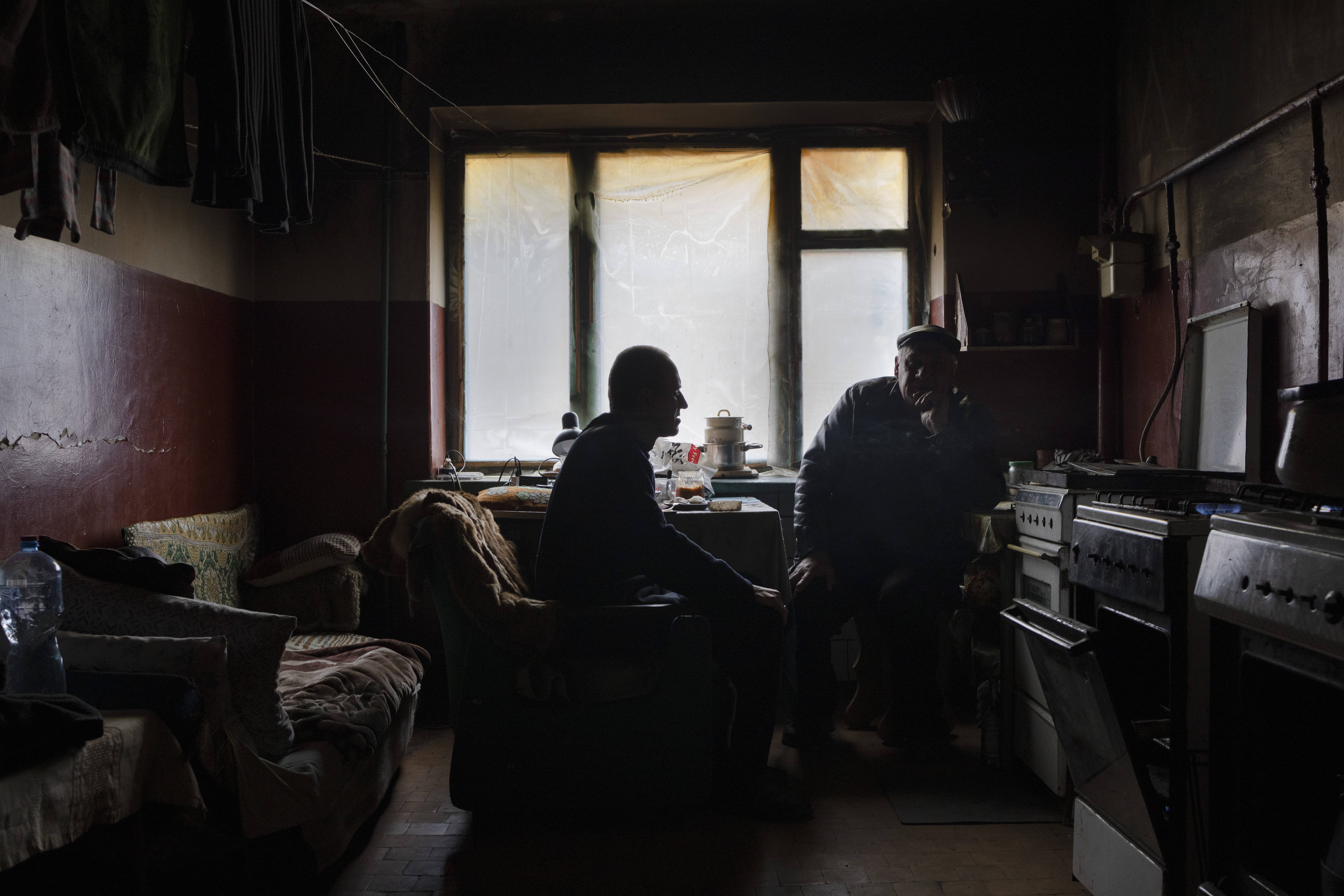 Friends Dmytro and Viktor in a communal kitchen of a damaged apartment building in Izium. The kitchen now served as a home for Dmytro, as most of the residents of the building had fled. He heats the space with the three gas stoves and sleeps on the corner of the room. Broken window has been fixed with a plastic cover. Dmytro and his friend Viktor were sharing a meal and watching Russian tv-series from a phone placed on the table. Despite the war they have not started to boycott Russian culture. They also reminded that they have been under crossfire, suffering from bombardment coming from both sides. 
