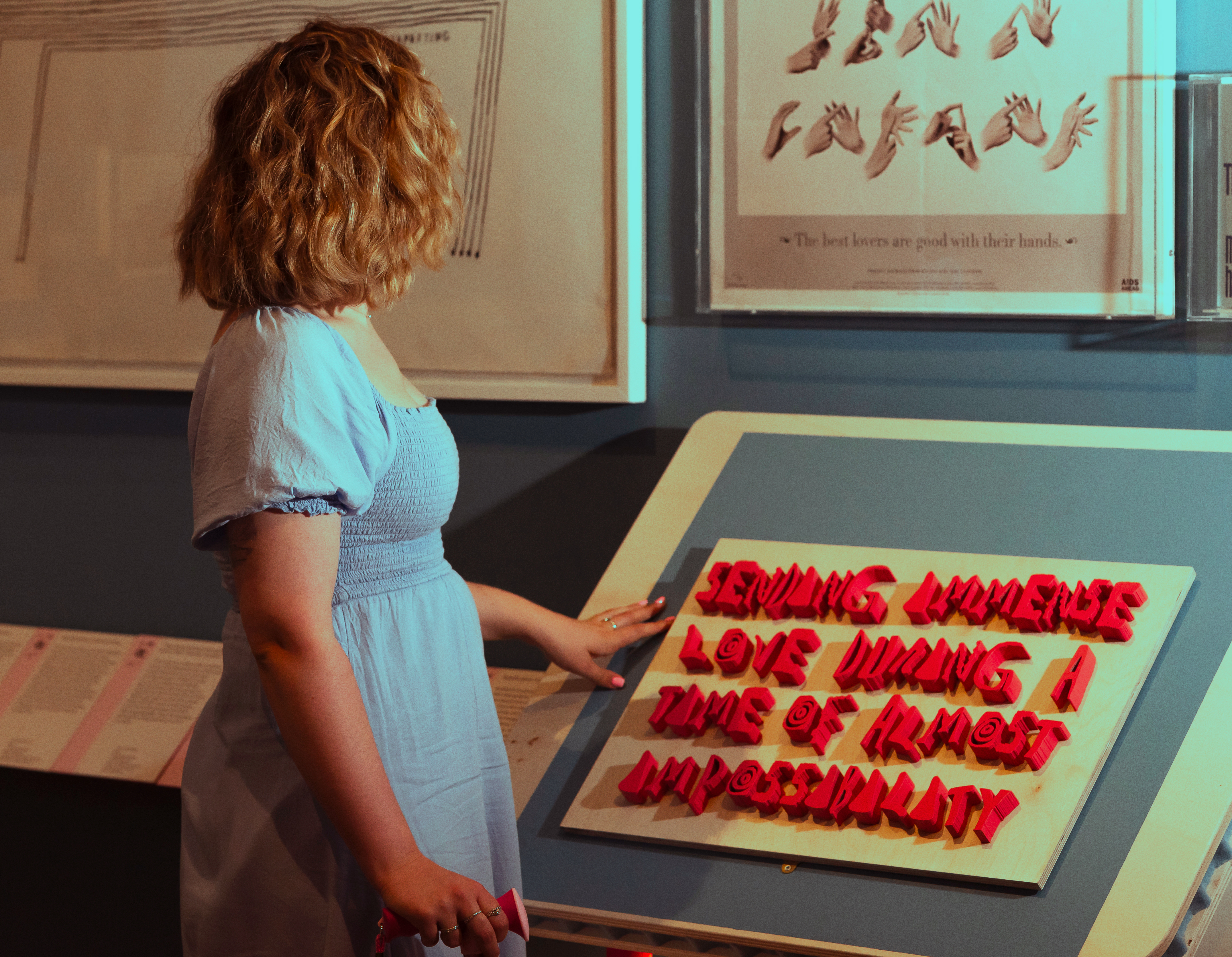 504 Font by Emily Sara (Installation shot of a sculpture by Emily Sara at the Design and Disability show, Victoria & Albert Museum, South Kensington. It is a 3D printed version of the 504 Font with touchable pink text that reads: Sending immense love during a time of almost impossibility. An individual with a blue dress and a cane is looking at the work. Photo by Isobel Greenhalgh.) 