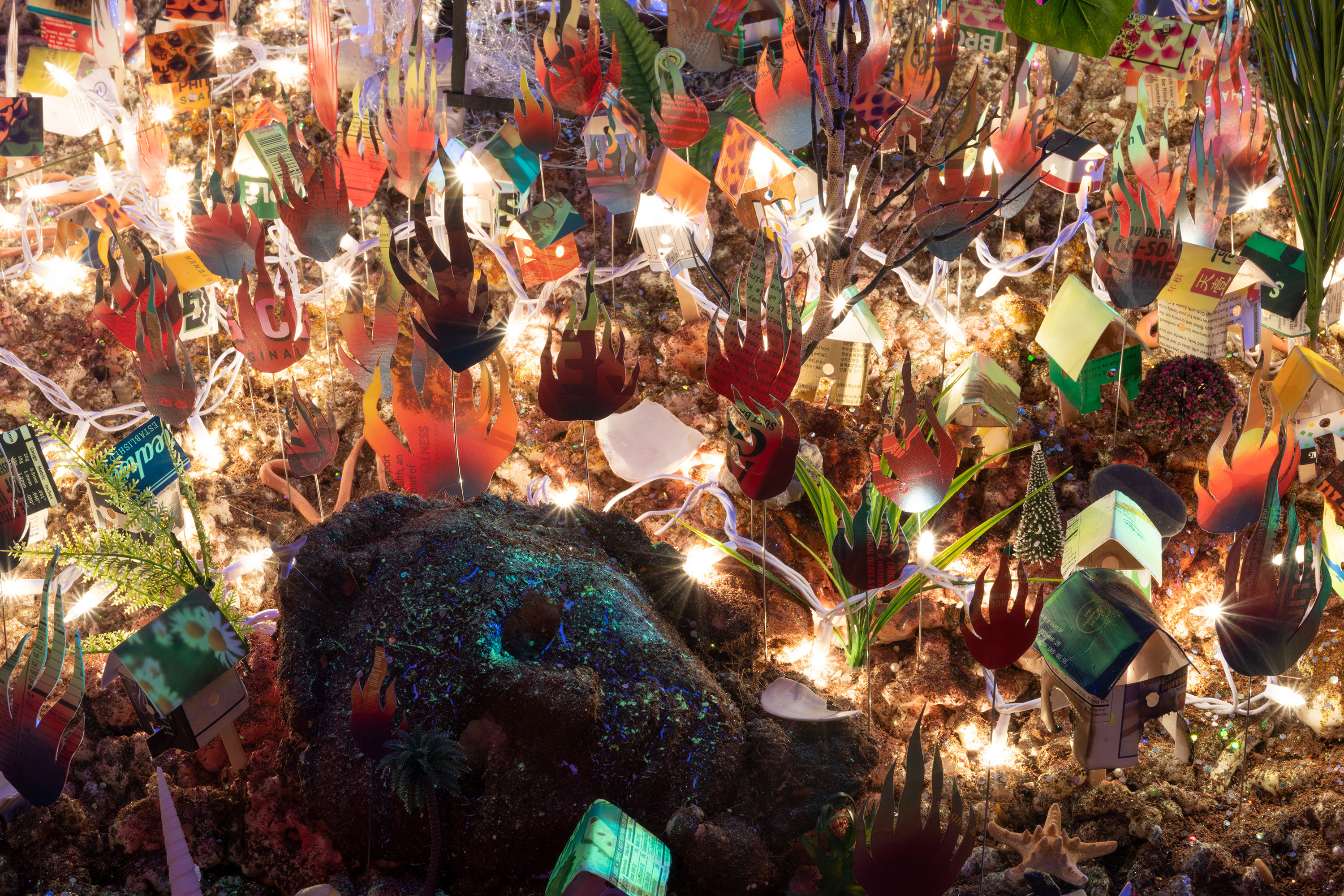 Orlando Estrada, Tiered Panorama, (detail) 2021, coconut fiber, expanding foam, glitter, artificial pearls, 3D printed PLA filament, sea shells, stone, artificial foliage, cell phones, Swarovski crystal, bone, consumer waste, found objects, light string, LED light, motor, UV pigment, acrylic, adhesive, PVC and Wire Armature 60” x 120” x 120”. Illuminated by strings of lights within the foreground, miniature houses made from consumer waste stand on stilts alongside a large foam head and various plants, bushels of fire surround them.