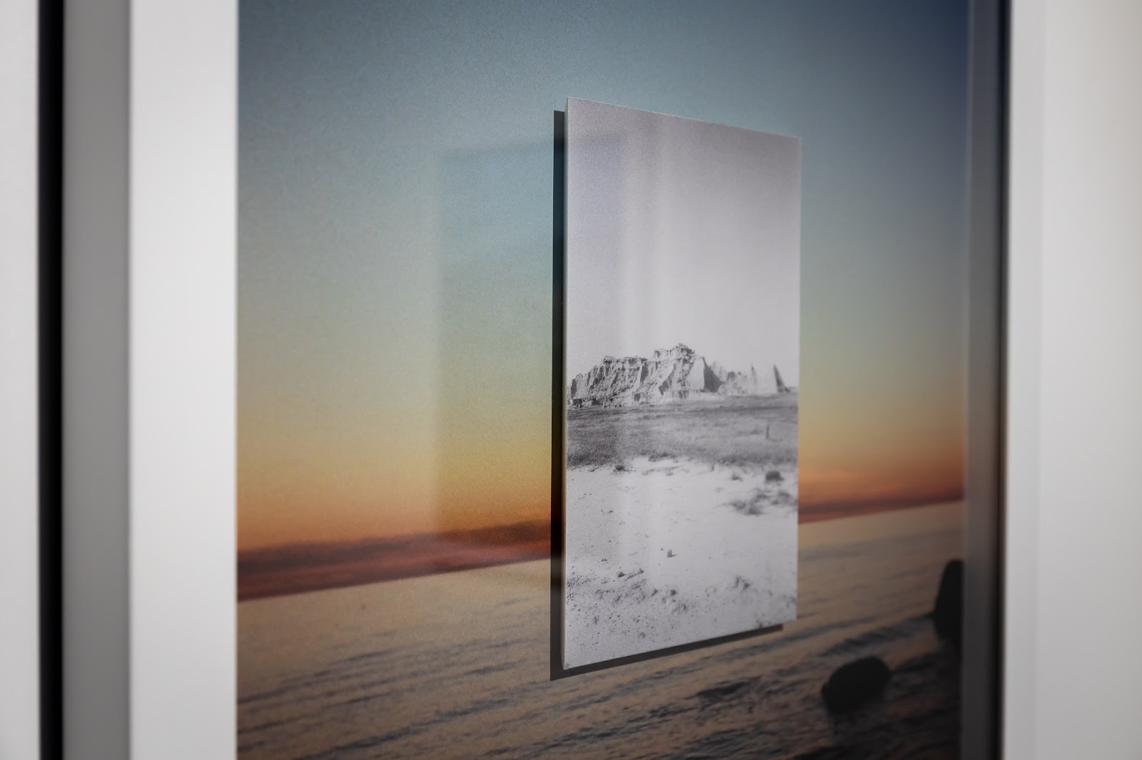 A close up of a color photograph with a white frame, depicting a dimly lit seascape just after sunset. There are dark blues and greens and reds and oranges. Superimposed in the center of this image is a smaller, black and white photograph of a desert landscape.