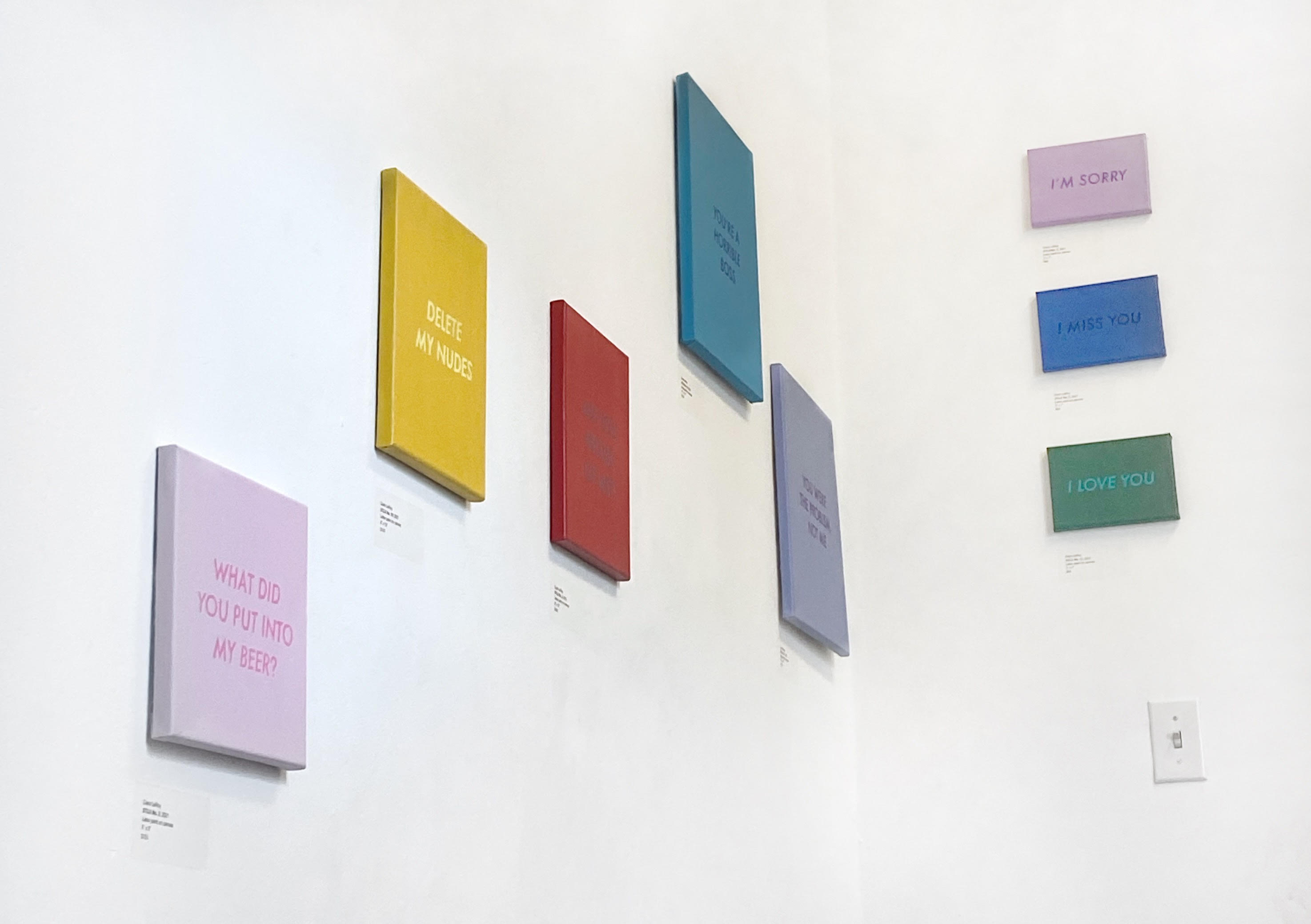 In the corner of two white walls hang 8 embroidery paintings, 5 on the left in a loose stair step pattern, and 3 smaller on the right vertically, in a line. They show text of a solid color on a background of a contrasting solid color, like lavender, yellow, blue, green, and red. One of the nearer texts reads, “Delete my nudes” and the farthest three read “I’m sorry,” “ I miss you,” and “I love you.”