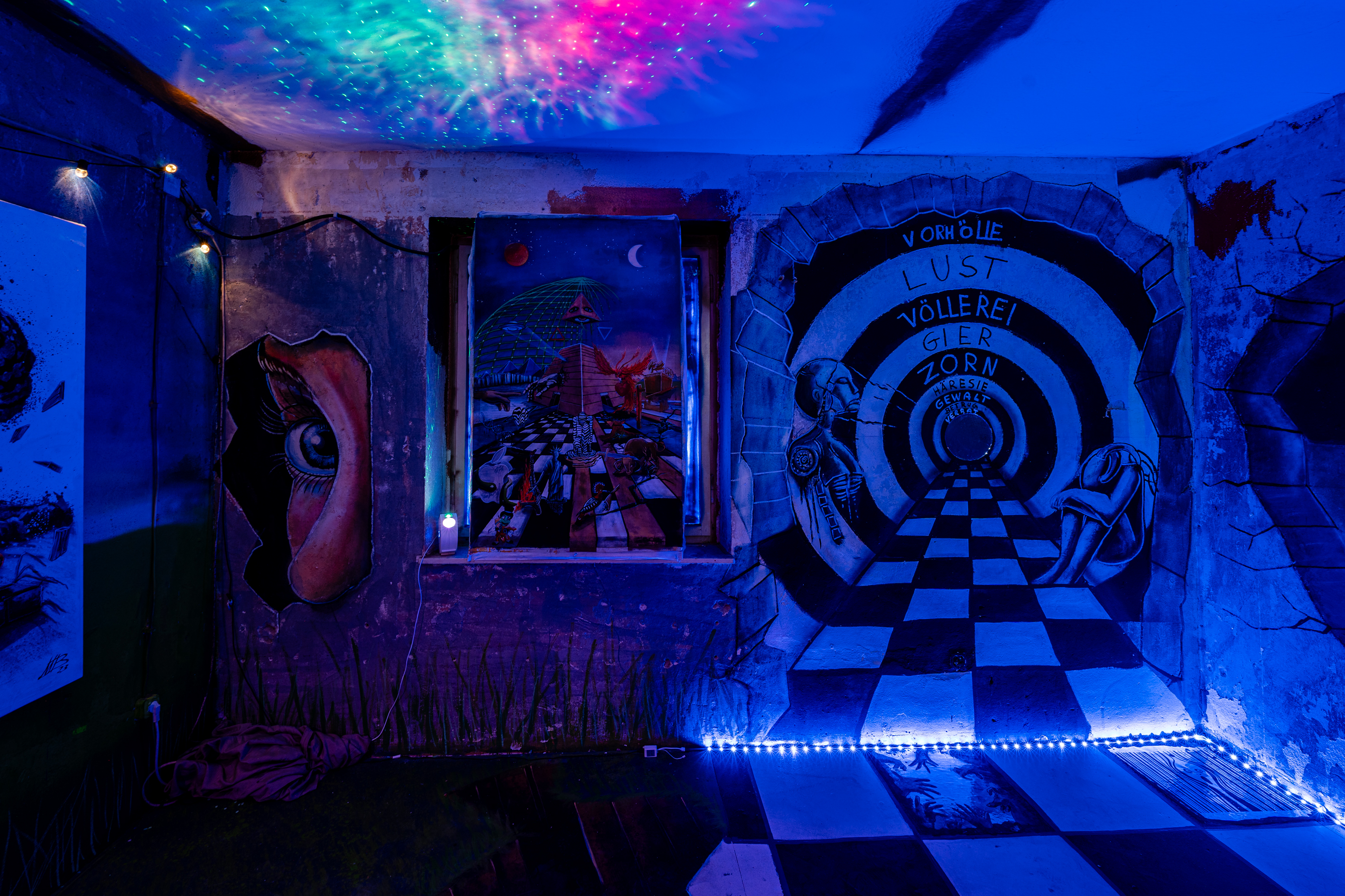 Bild 1 Eyes Wide Shut: Ein abgedunkelter, blau ausgeleuchteter Raum mit Wandmalereien. Diese spielen mit optischen T&auml;uschungen. Der Boden des Raumes wurde in der rechten H&auml;lfte mit schwarz-wei&szlig;em Schachbrettmuster bemalt. Dieses verl&auml;uft an der Wand weiter und wird schmaler. 