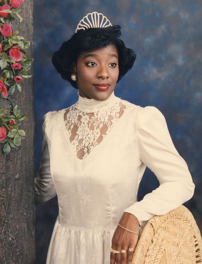 Image: A color portrait of Ms. J. Chantele Williams who was "Miss Philander Smith College" during 1992-1993. She wears a white dress, crown, and rings on all of her visible left fingers. She stands next to a faux tree bark with flowers draped at its top, with her arm propped on a wicker chair. Philander Smith College Digital Archive. Photographer: Unknown.