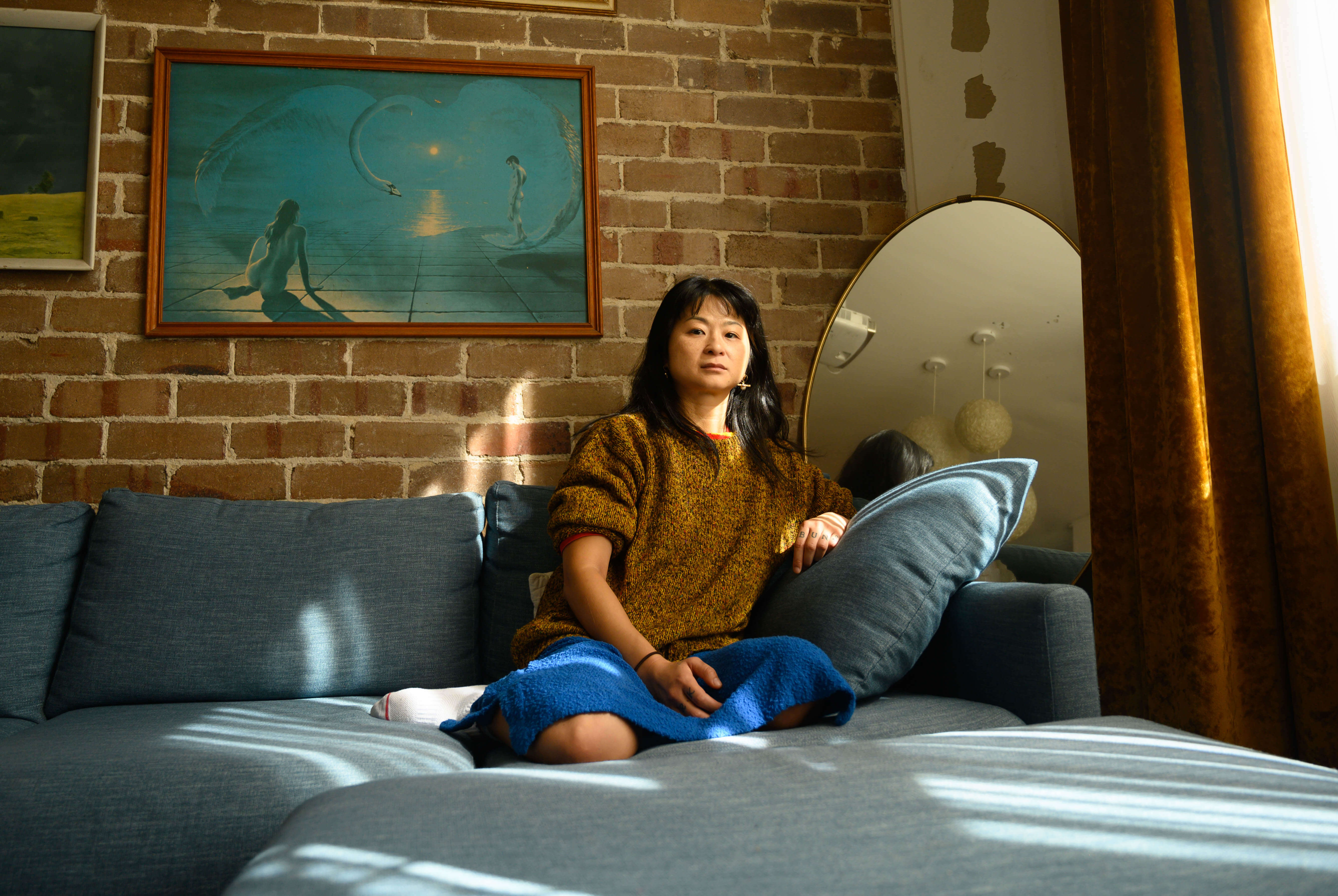 Joy, a woman of East Asian descent sits on her blue sofa. She gazes directly at the camera from a higher vantage point.