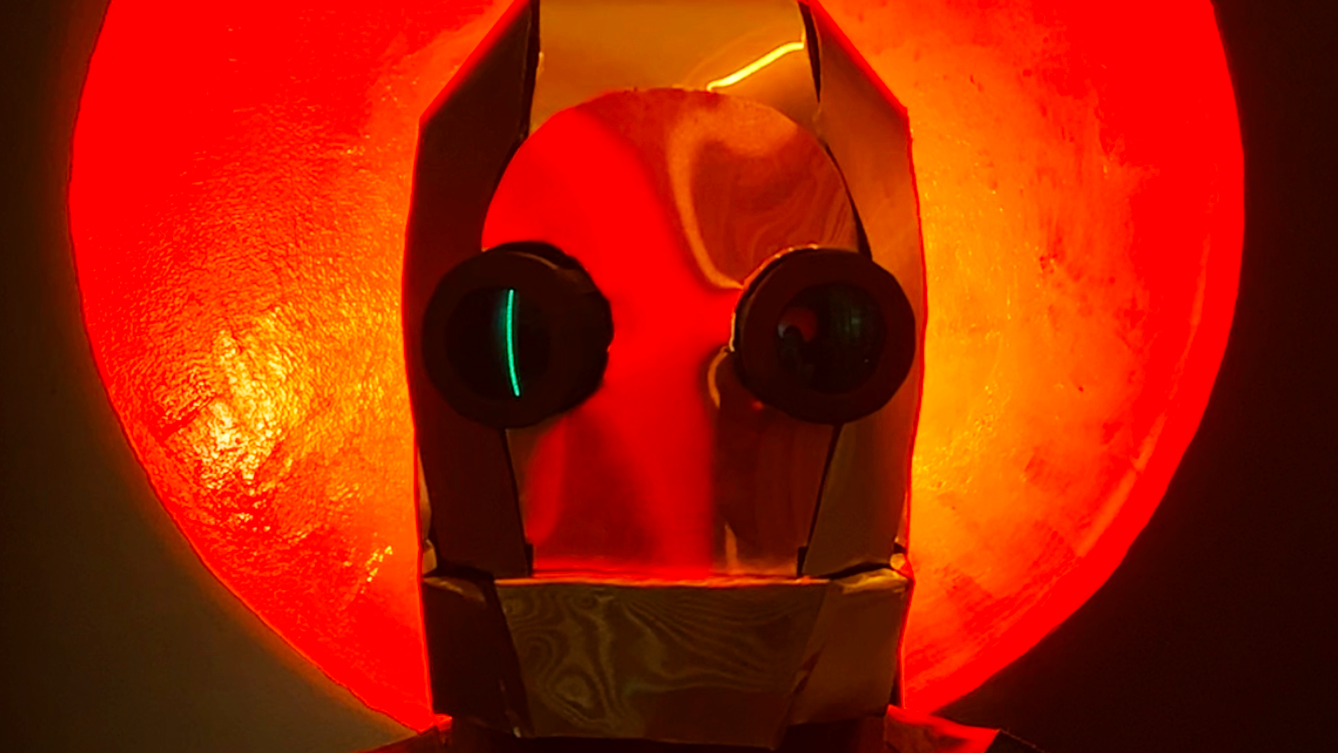 A metallic robot head lit with orange light, sitting in front of a large, orange glowing circle on a dark green wall.