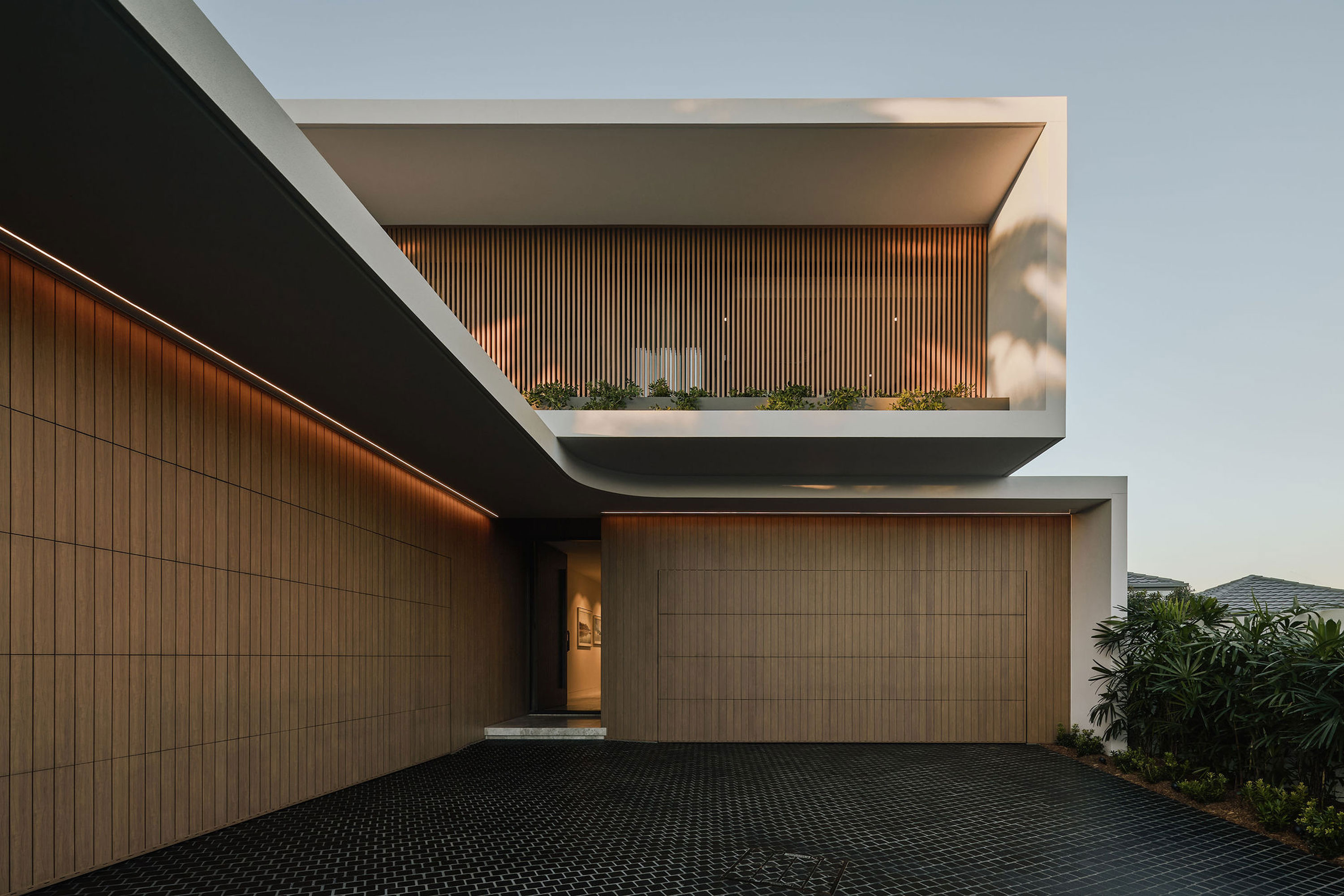 Close-up view of modern minimalist house exterior with vertical wooden slat cladding, concealed garage door, black tiled driveway, integrated LED strip lighting, and upper balcony with greenery and sleek white frame.