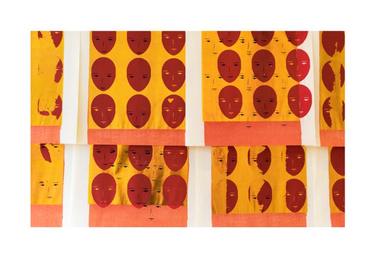 A series of red feminine faces printed on gold paper with a white border. The features on the faces are disorganized, obscured, and appear as if misprinted.  