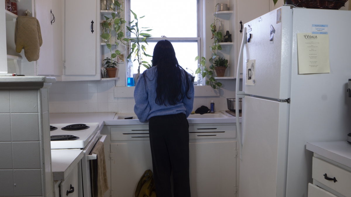 Woman back in her own apartment's kitchen, which is noticeably bare compared to her family's kitchen; woman washing dishes