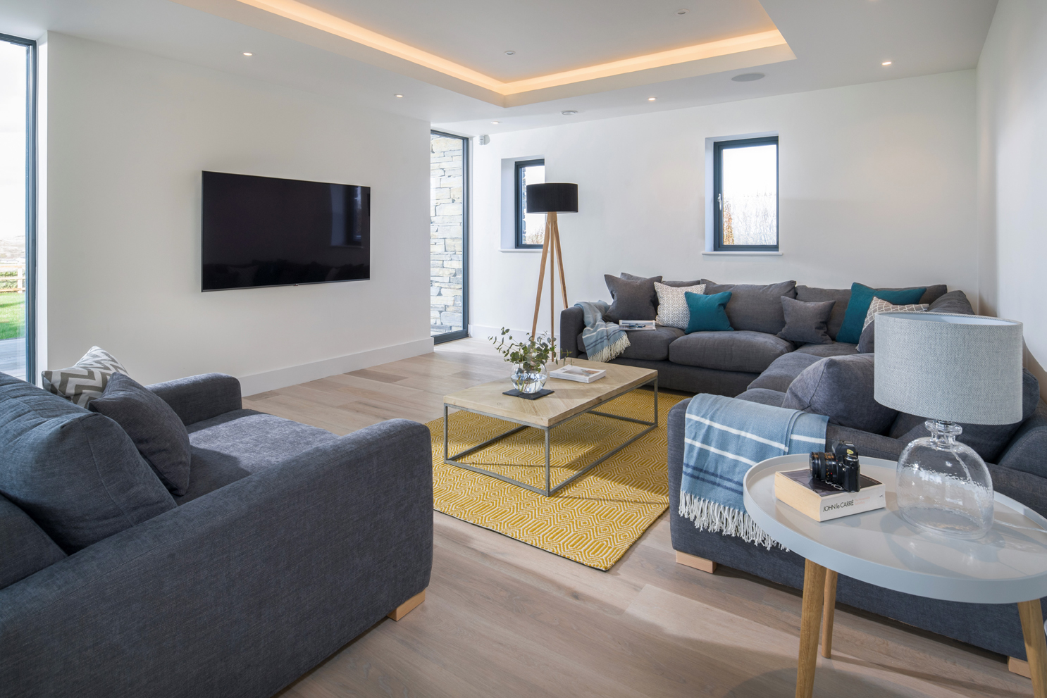 Modern lounge with grey furnishings, mustard yellow rug and coffee and side table. Recessed ceiling lighting. TV on wall.