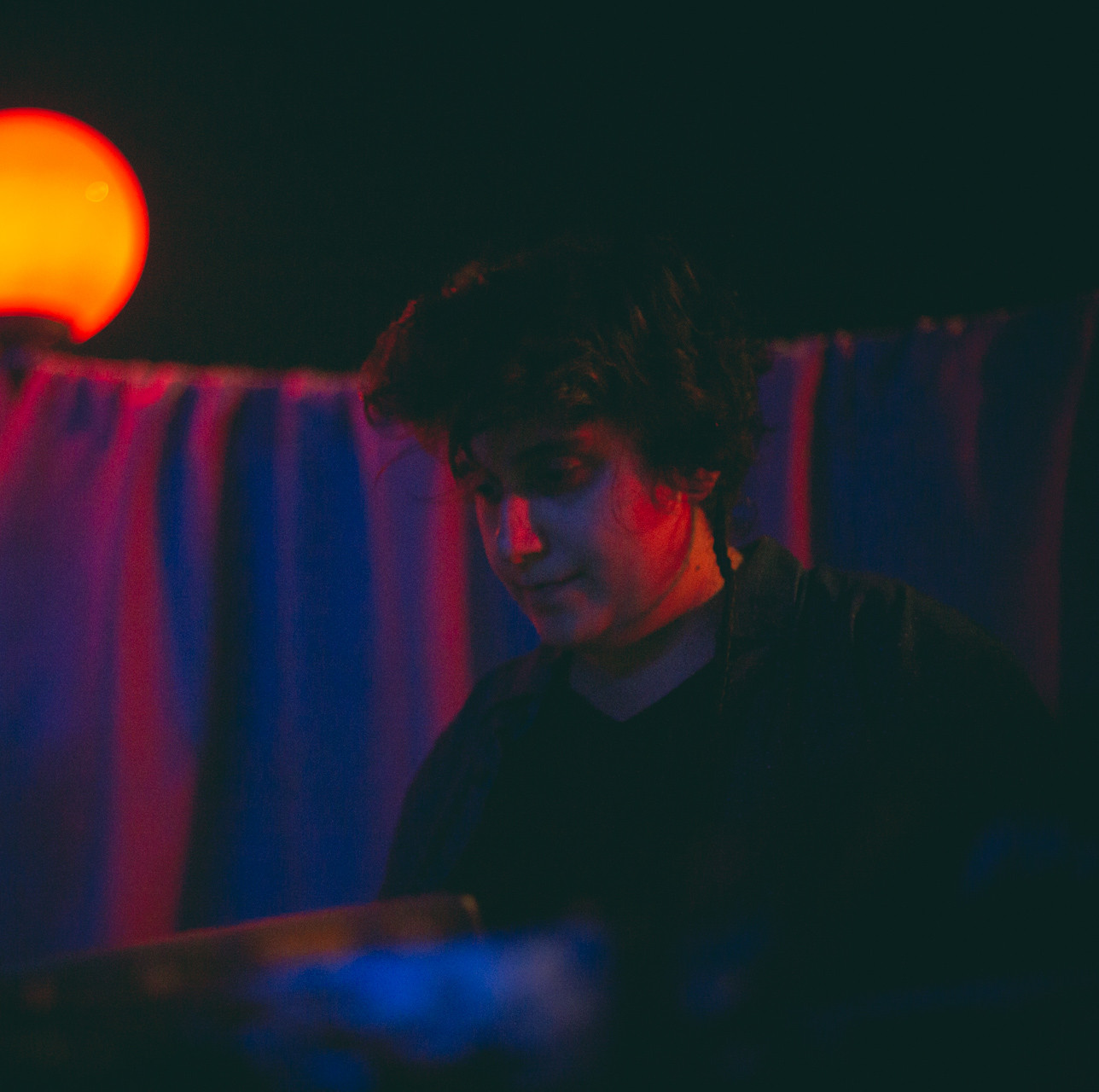 [IMAGE DESCRIPTION]: Portrait of Melis, in red and blue lights at a party, with a wavy fabrics backdrop.
