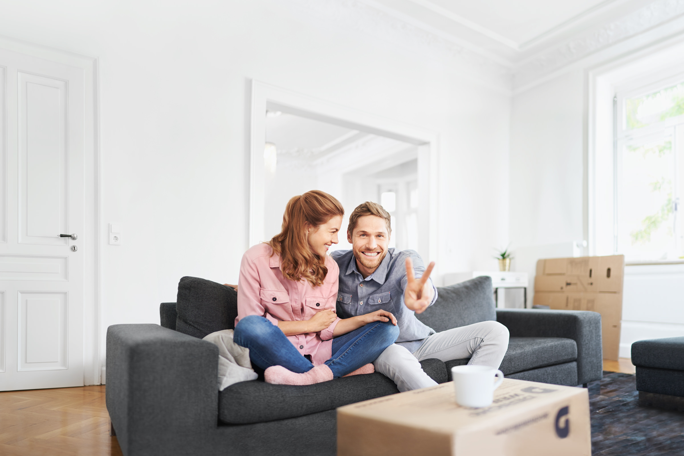 Portrait eines Paerchens, sitzend auf einer Couch in einem Wohnzimmer. Der Mann zeigt das Victory-Zeichen in die Kamera. Umzugskisten deuten auf einen Umzug hin.