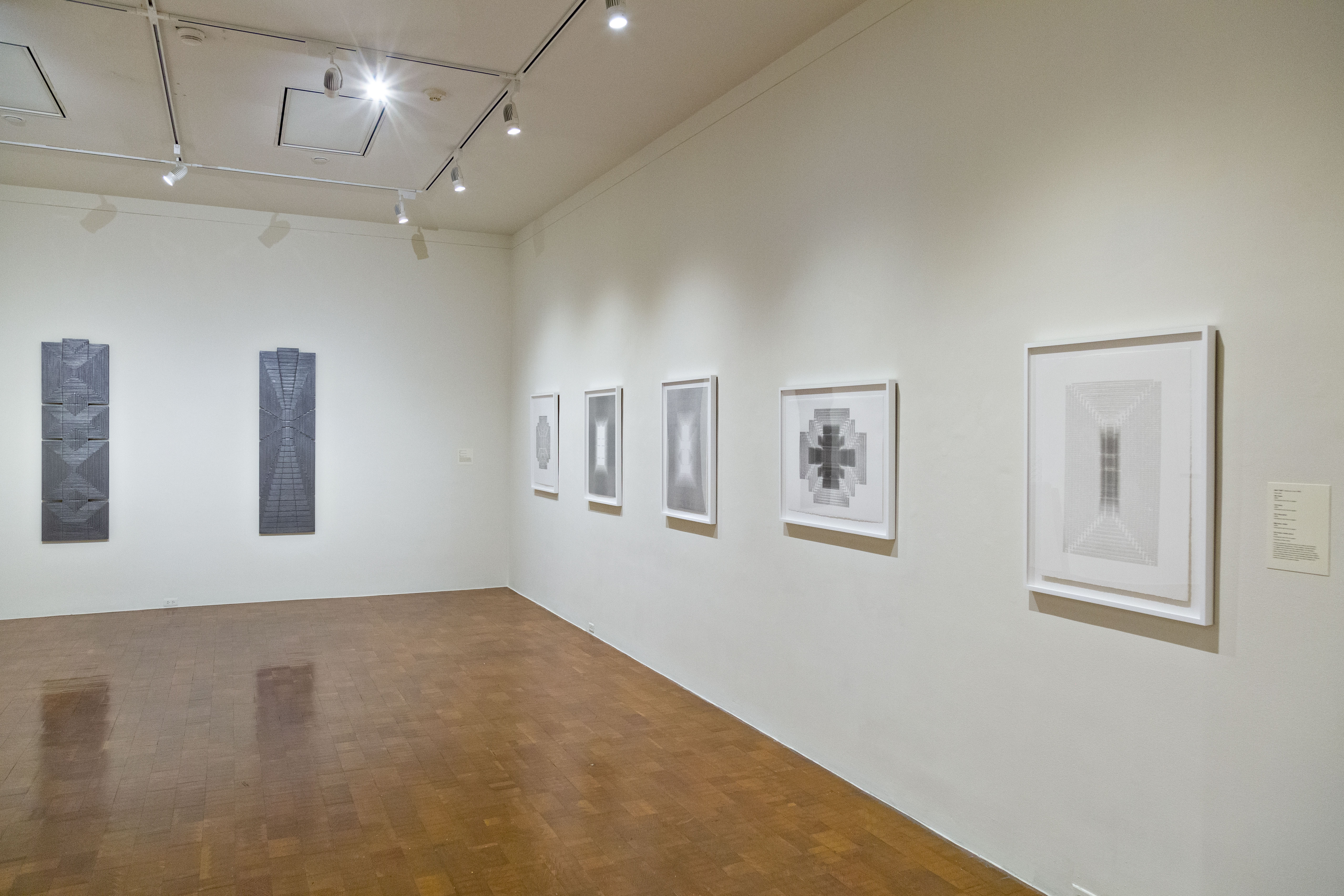 View of a sparse, white wall museum space with shiny wood floors and track lighting overhead. On the back/left wall hangs two long, rectangular dark grey and blue works. On the right wall, closer, are more works, lighter in color and framed. They consist of tight geometric images.