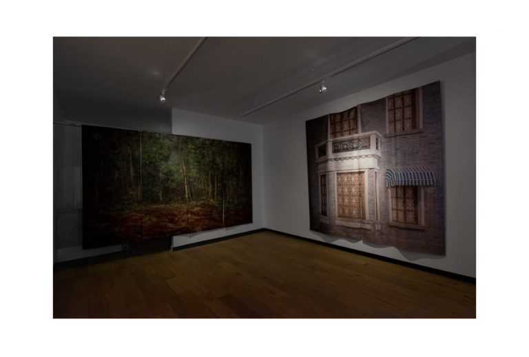 A photo of the gallery exhibition. A painting of a forest is hung on the back wall. A printed photograph of a building facade juts out from the right wall as a 3D wall installation.