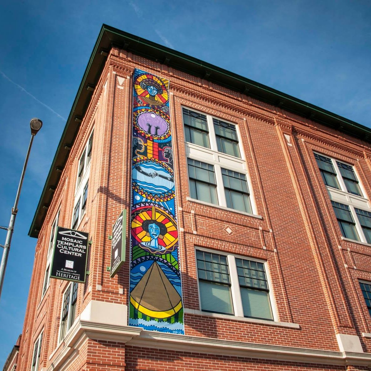 Image: Mural created by Perrion Hurd on the exterior of the Mosaic Templars Cultural Center. The photo is looking up at an outer corner of the brick building with a tall, colorful mural in view, all against a blue sky.
