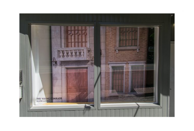 A photograph of the storefront of the gallery. The vinyl on the window reads: "The Young Comrade, Alvin Luong". Inside the front windows we see a large printed photograph of a facade of a building from the movie "The Young Comrade". This printed photograph takes up the whole window space.